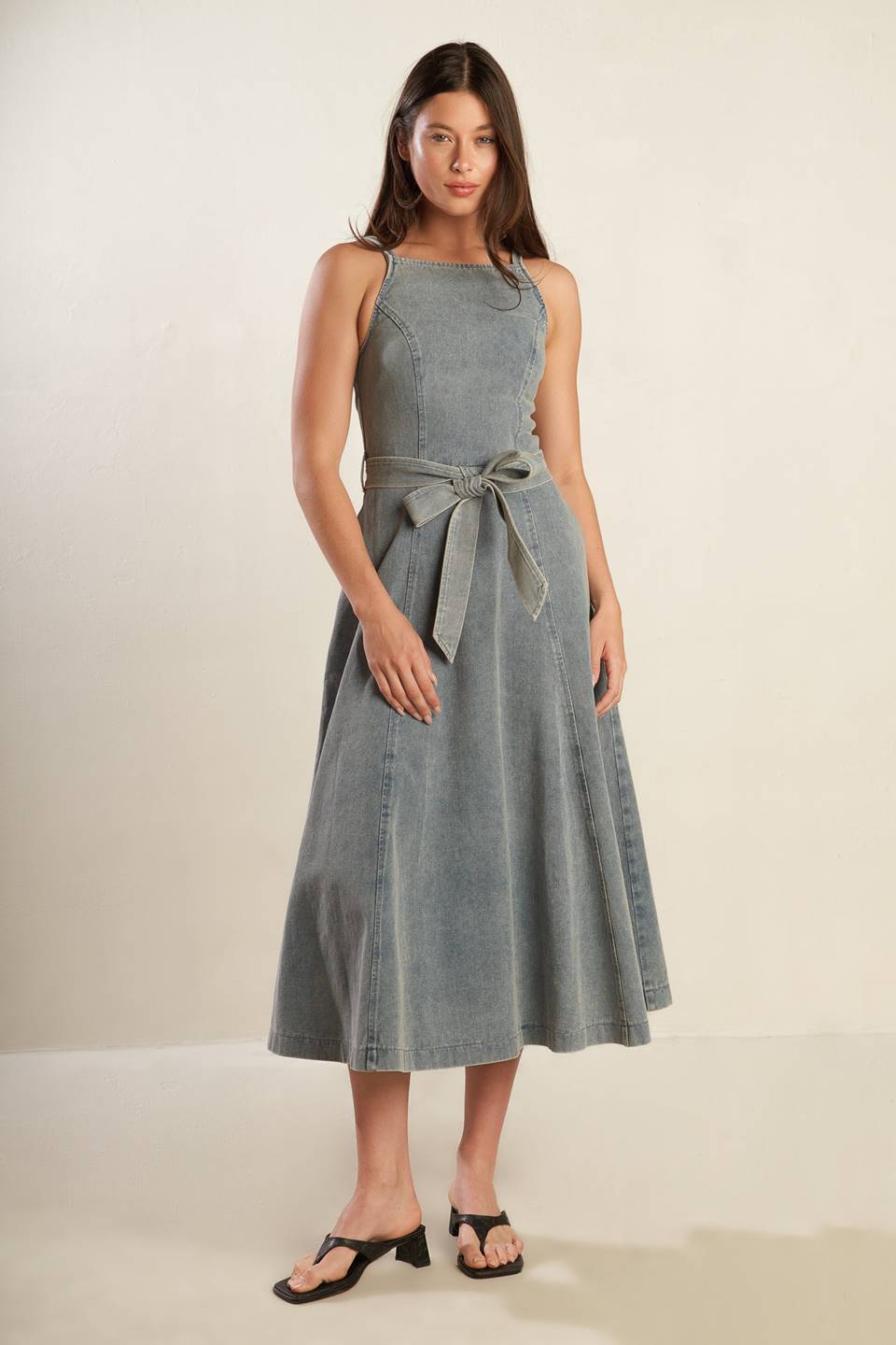 A washed denim midi dress featuring straight neckline, straps, circle skirt, self sash tie and back zipper closure.