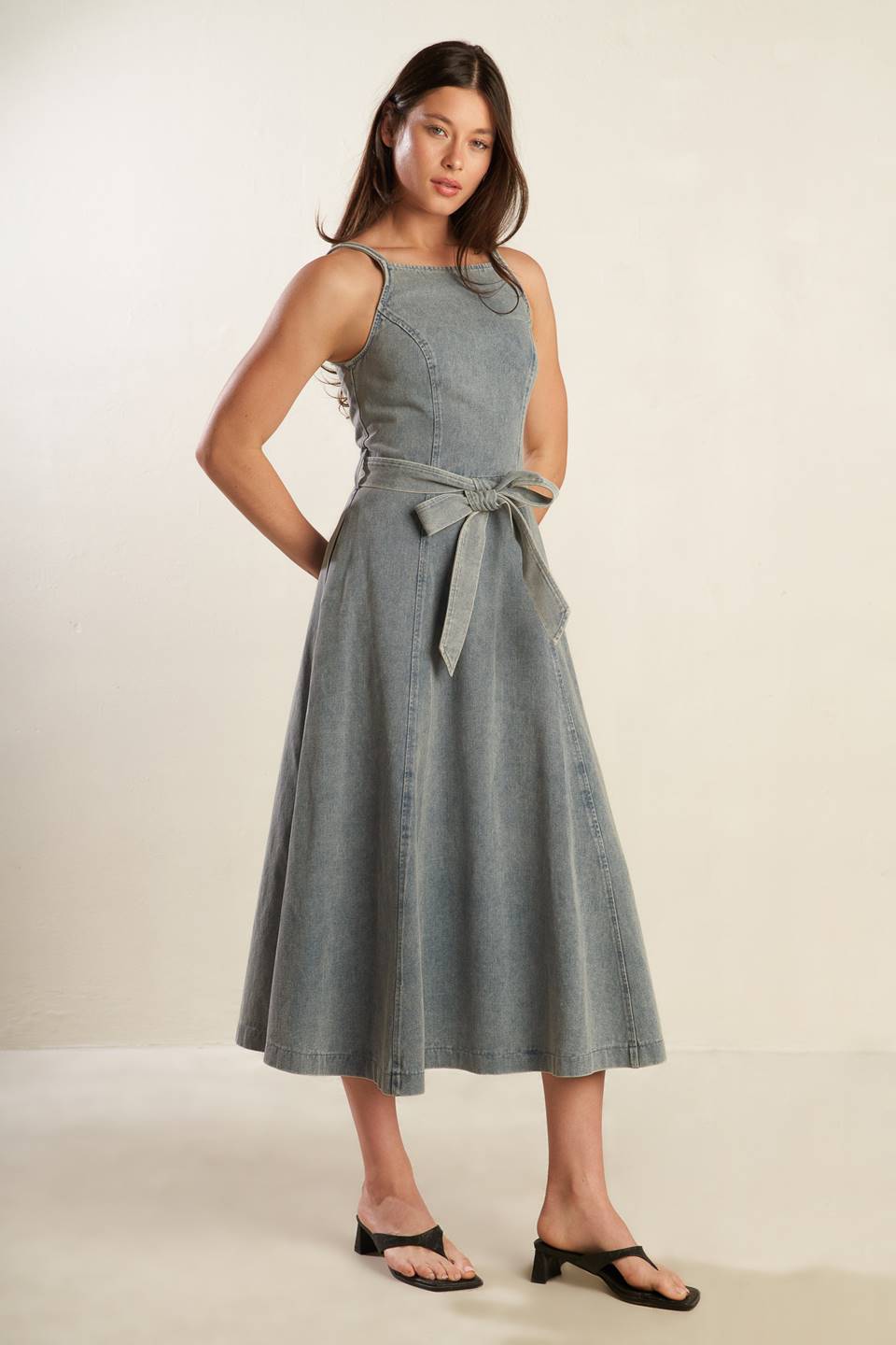 A washed denim midi dress featuring straight neckline, straps, circle skirt, self sash tie and back zipper closure.