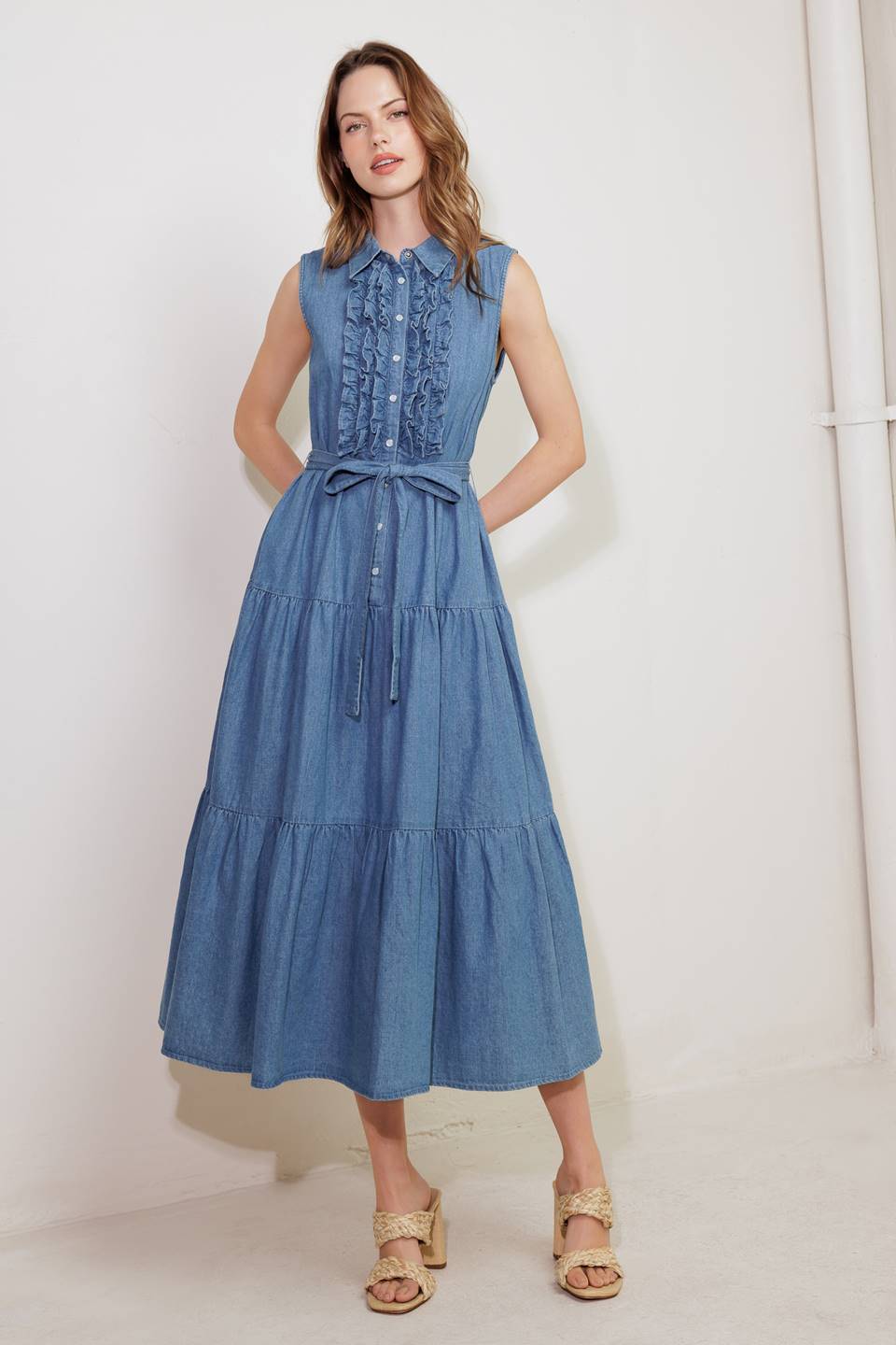 A washed denim midi dress featuring collar, snap button down, ruffled front, sleeveless, self sash tie and tiered skirt
