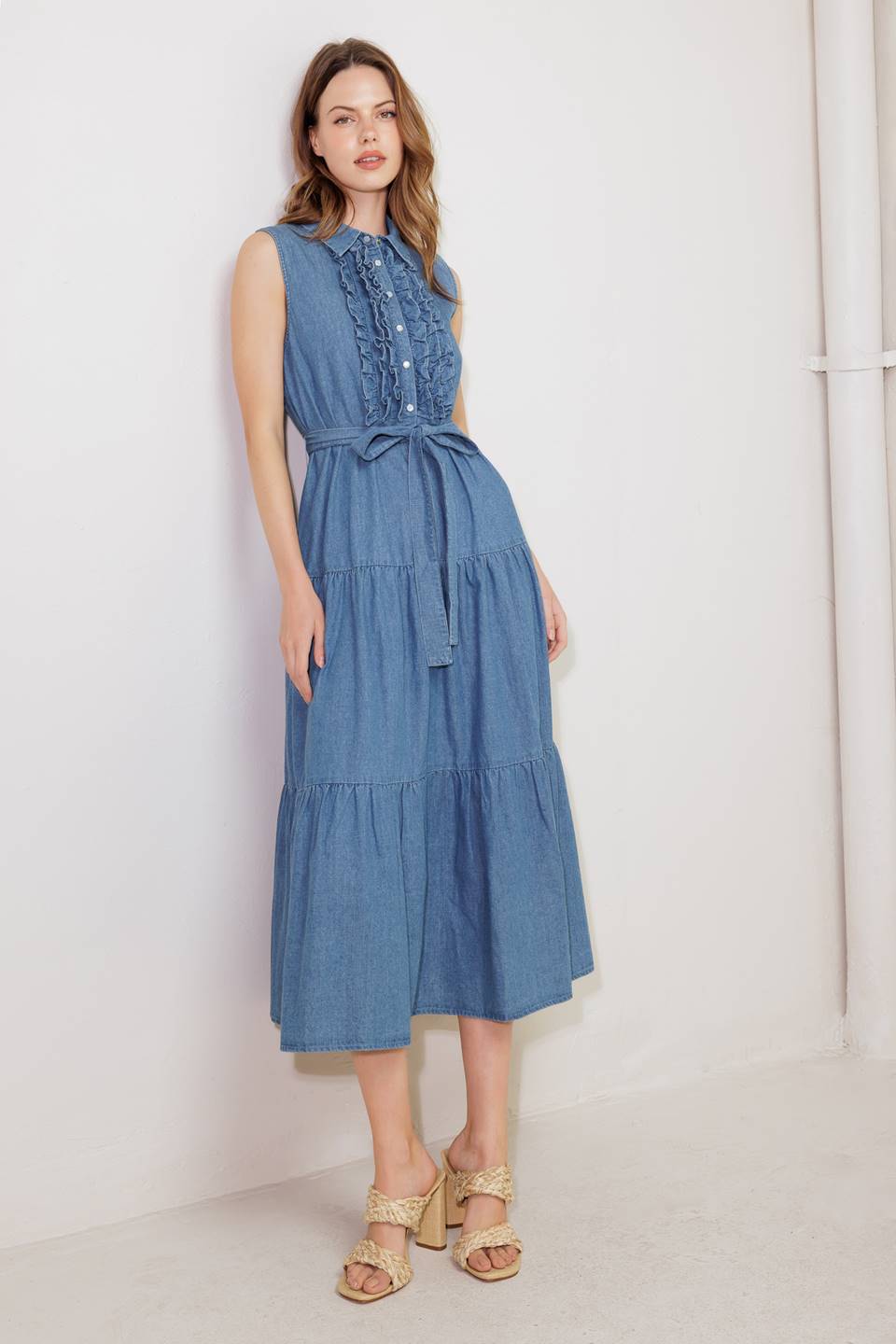 A washed denim midi dress featuring collar, snap button down, ruffled front, sleeveless, self sash tie and tiered skirt