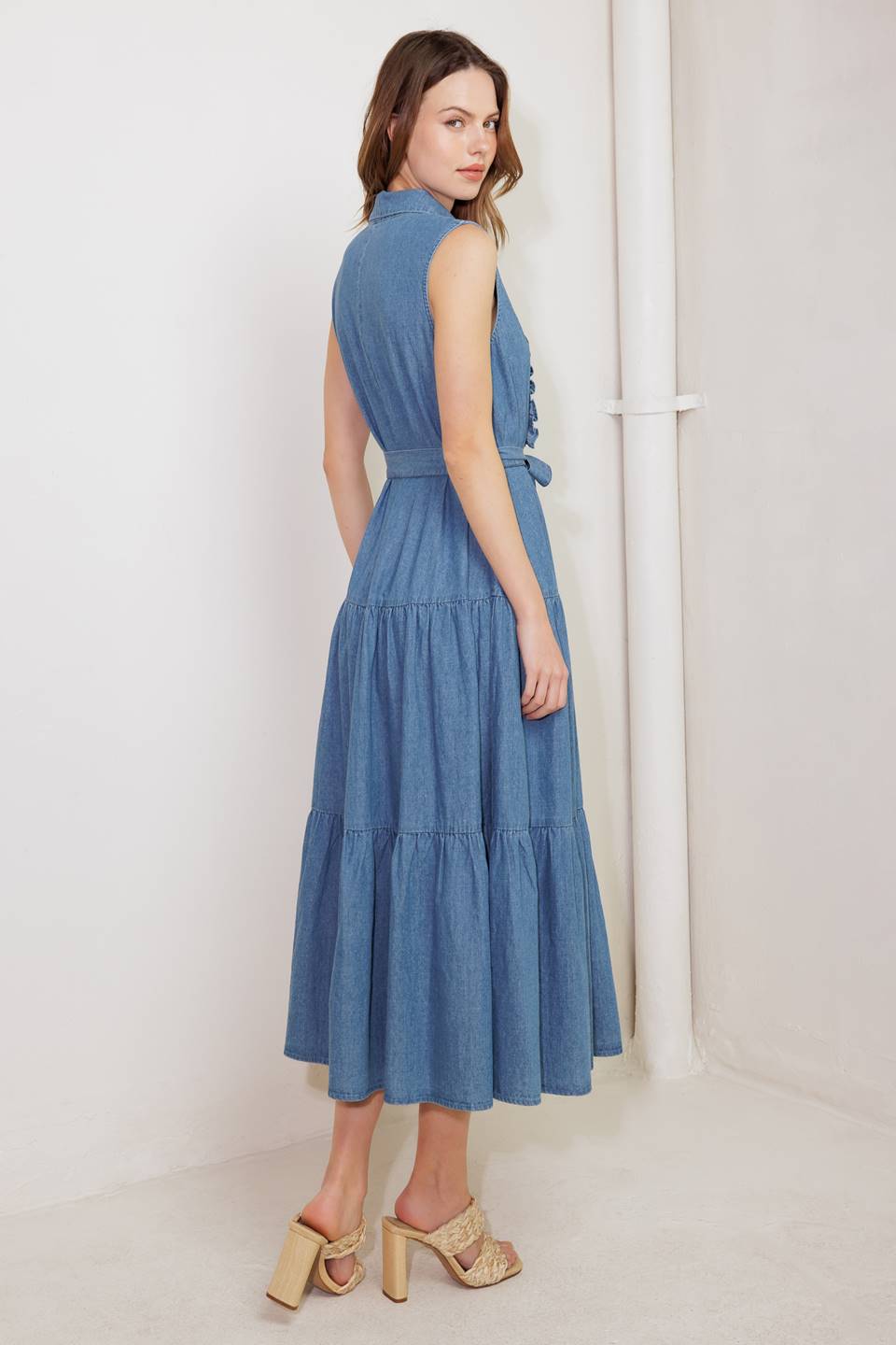 A washed denim midi dress featuring collar, snap button down, ruffled front, sleeveless, self sash tie and tiered skirt
