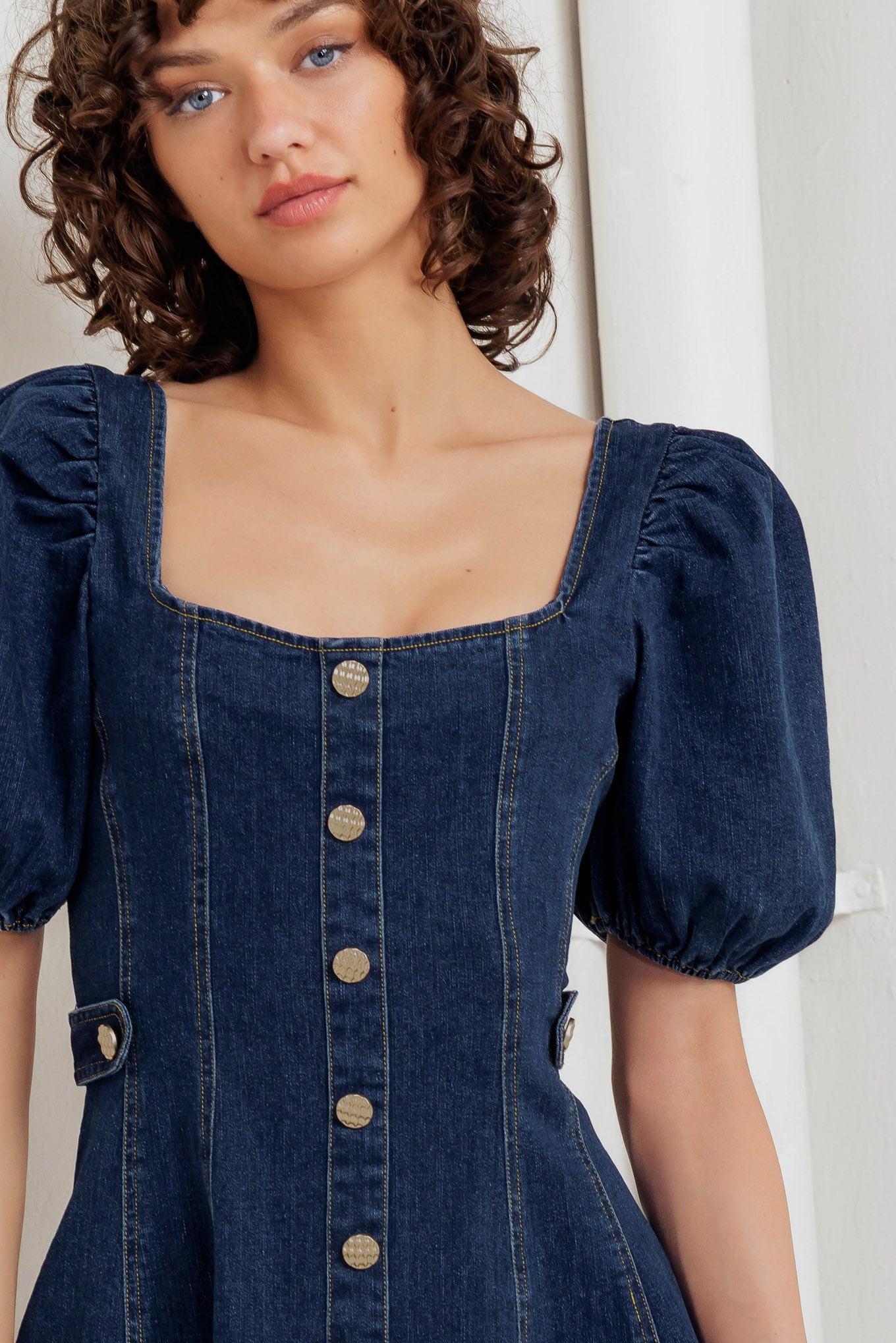 A washed denim mini dress featuring square neckline, short puff sleeve, button down detail, circle skirt and back zipper closure