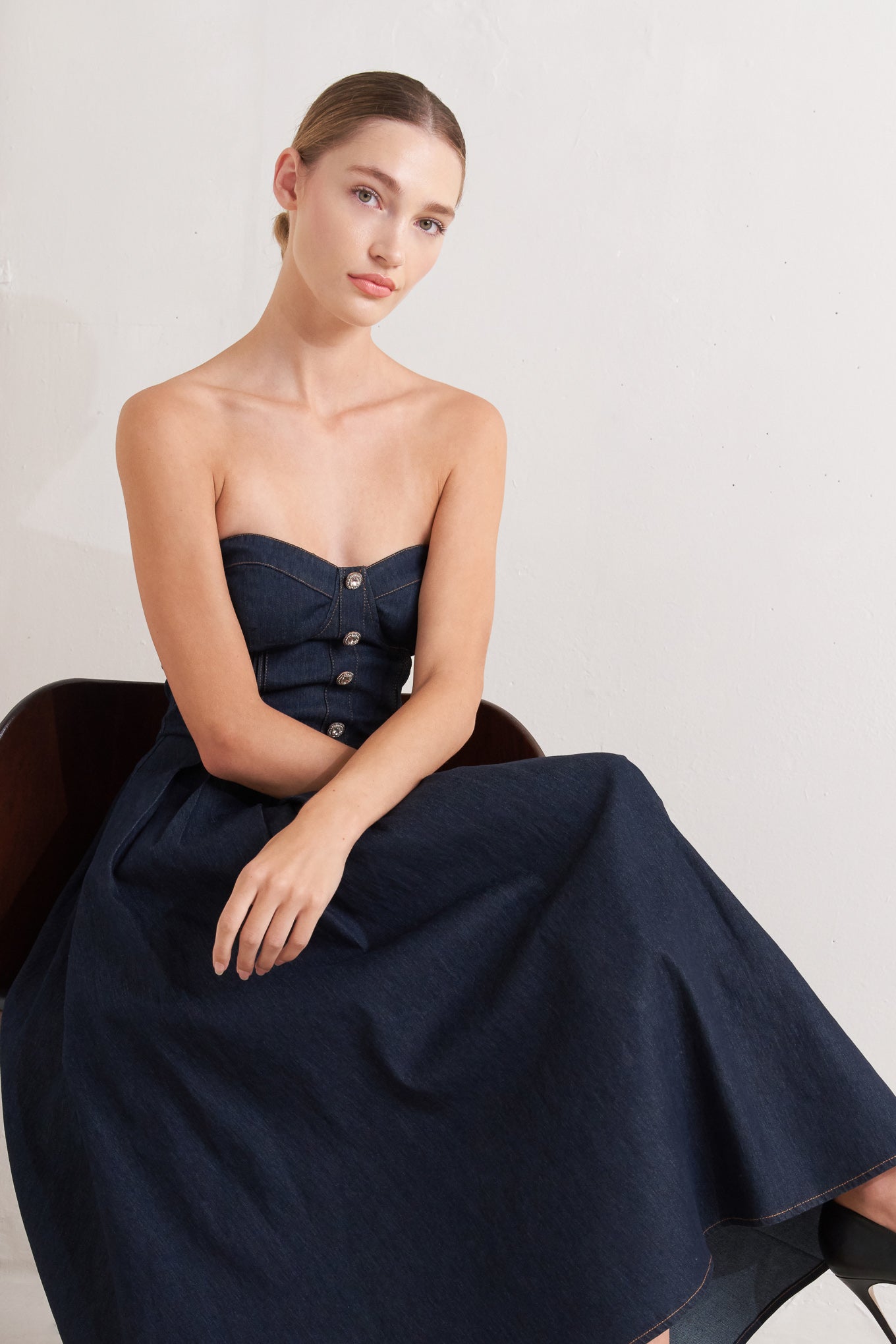 A denim midi dress featuring strapless sweetheart neckline, bustier bodice with button detail, basque waist, full skirt and back zipper closure