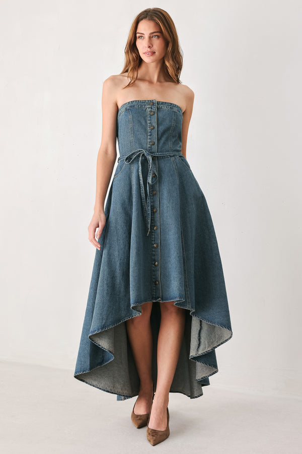A woman modeling a strapless high-low denim dress in a dusty blue wash. The dress features a buttoned bodice, a matching fabric tie belt at the waist, and a flared skirt with an asymmetrical high-low hem. | FREEMARKET
