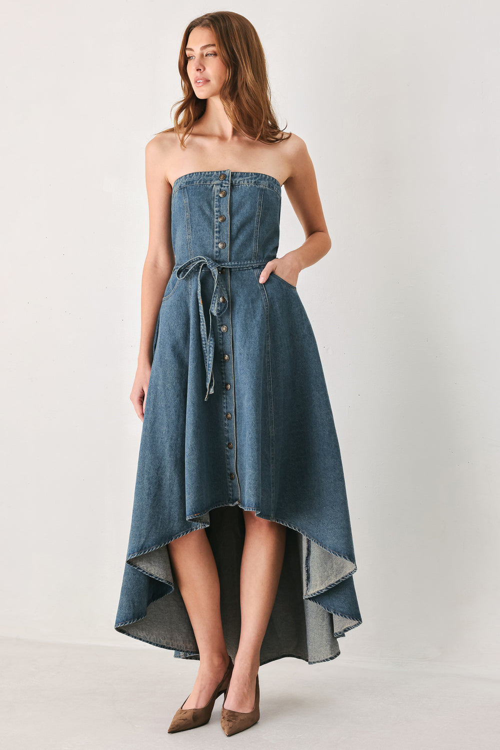 A woman modeling a strapless high-low denim dress in a dusty blue wash. The dress features a buttoned bodice, a matching fabric tie belt at the waist, and a flared skirt with an asymmetrical high-low hem. | FREEMARKET