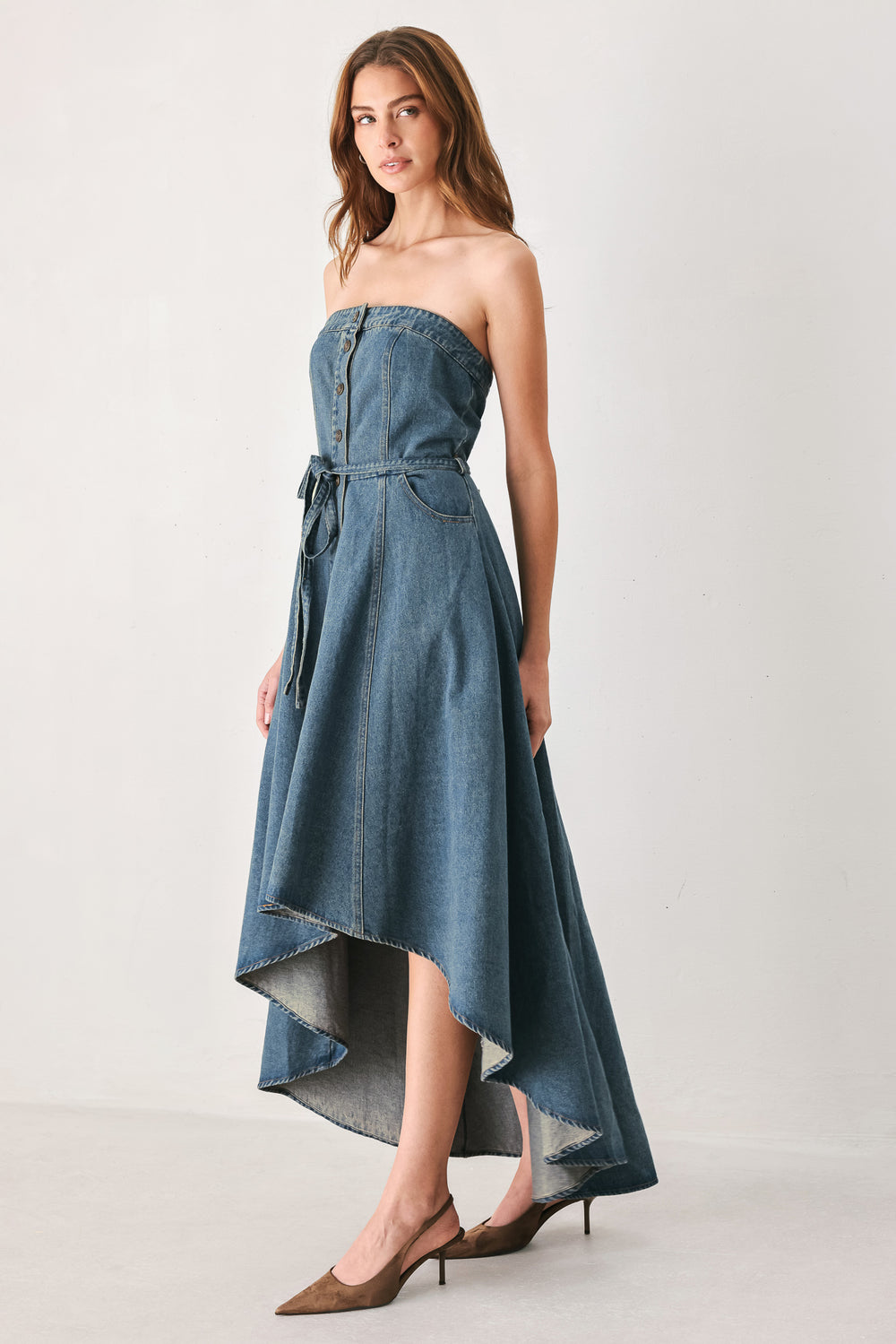 A woman modeling a strapless high-low denim dress in a dusty blue wash. The dress features a buttoned bodice, a matching fabric tie belt at the waist, and a flared skirt with an asymmetrical high-low hem. | FREEMARKET