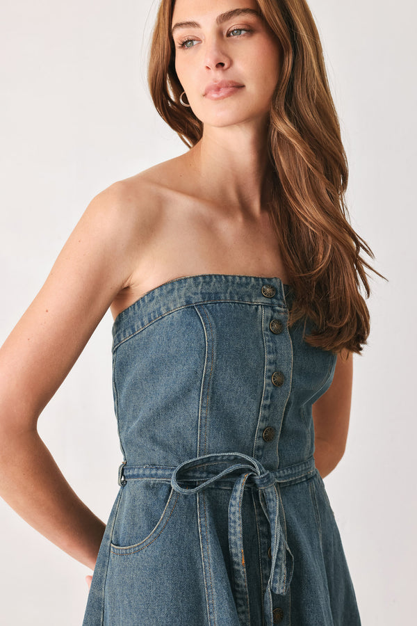 A woman modeling a strapless high-low denim dress in a dusty blue wash. The dress features a buttoned bodice, a matching fabric tie belt at the waist, and a flared skirt with an asymmetrical high-low hem. | FREEMARKET