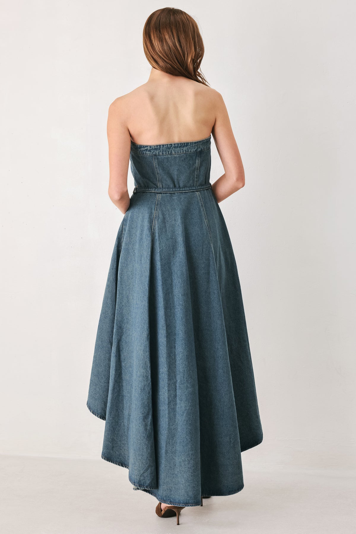 A woman modeling a strapless high-low denim dress in a dusty blue wash. The dress features a buttoned bodice, a matching fabric tie belt at the waist, and a flared skirt with an asymmetrical high-low hem. | FREEMARKET