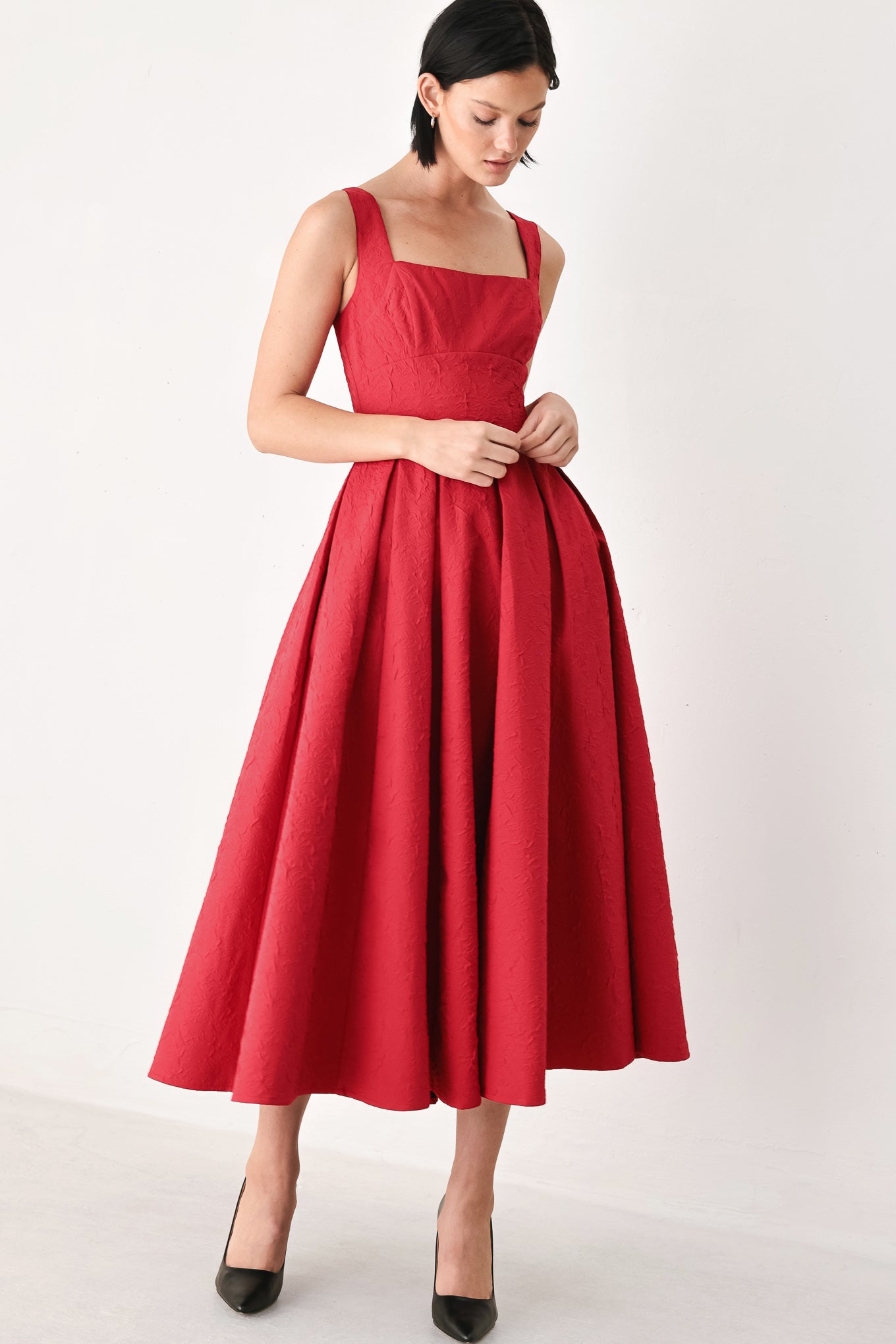 A woman modeling a sleeveless red midi dress with a square neckline, structured bodice, and full pleated skirt, paired with black pointed-toe heels against a plain light background.