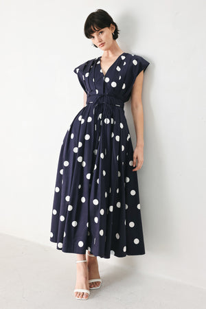 Woman wearing a navy blue midi dress with white polka dots featuring V neckline, short sleeve, front button, corset waist, double tie and full skirt | FREEMARKET