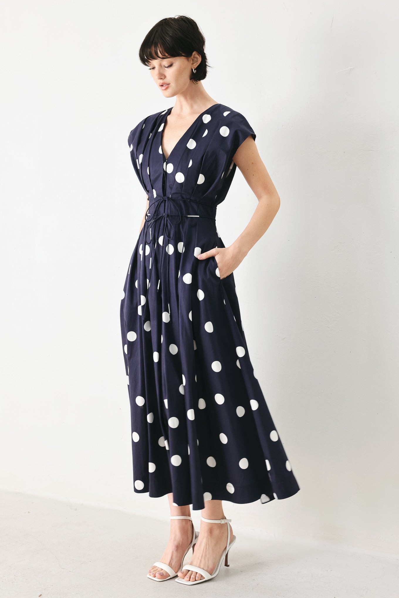 Woman wearing a navy blue midi dress with white polka dots featuring V neckline, short sleeve, front button, corset waist, double tie and full skirt | FREEMARKET
