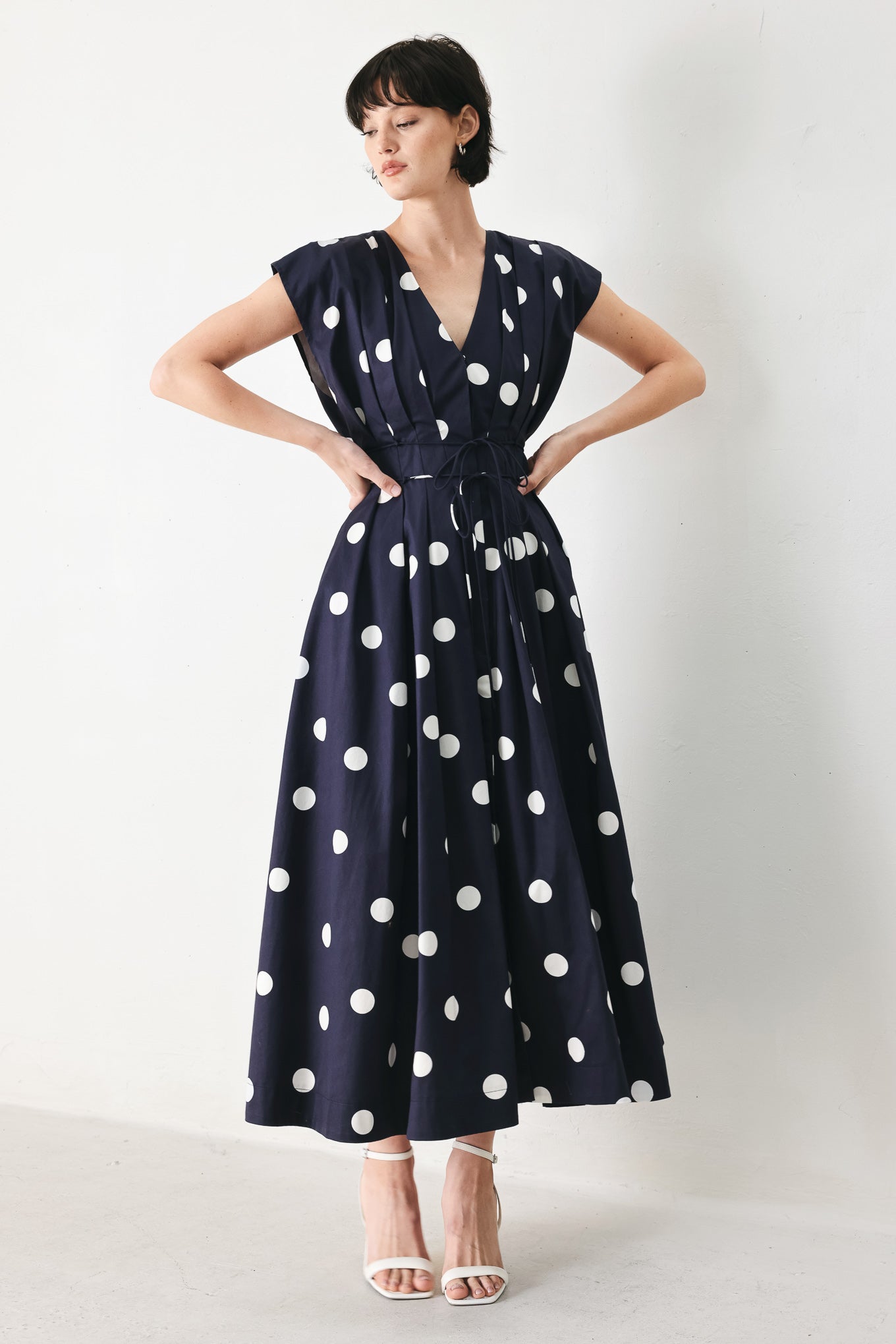 Woman wearing a navy blue midi dress with white polka dots featuring V neckline, short sleeve, front button, corset waist, double tie and full skirt | FREEMARKET