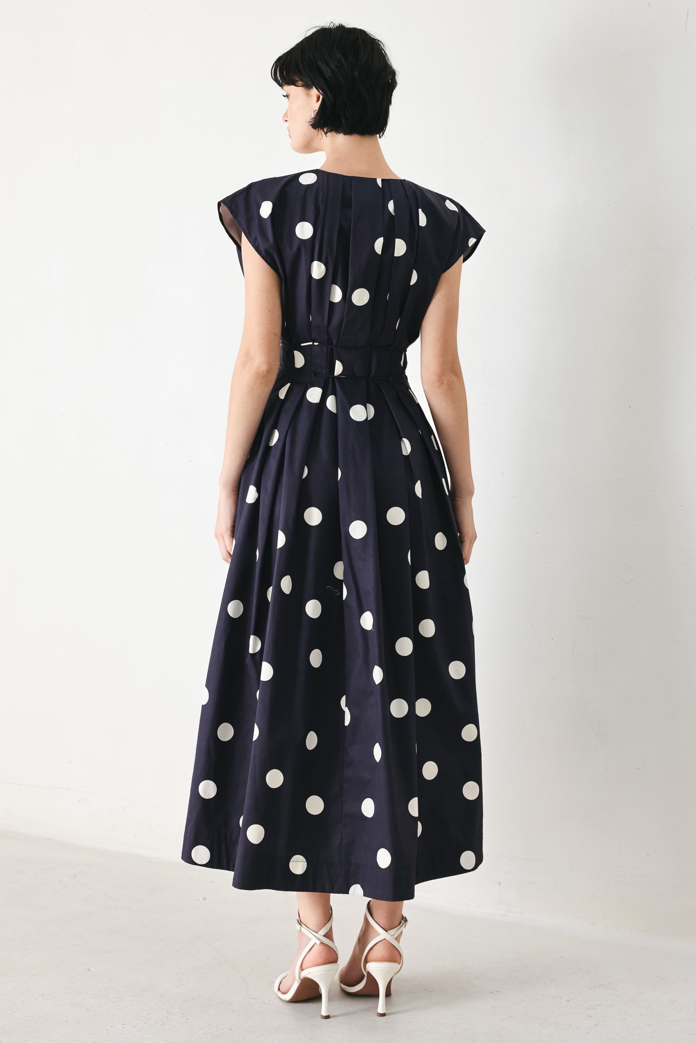 Woman wearing a navy blue midi dress with white polka dots featuring V neckline, short sleeve, front button, corset waist, double tie and full skirt | FREEMARKET