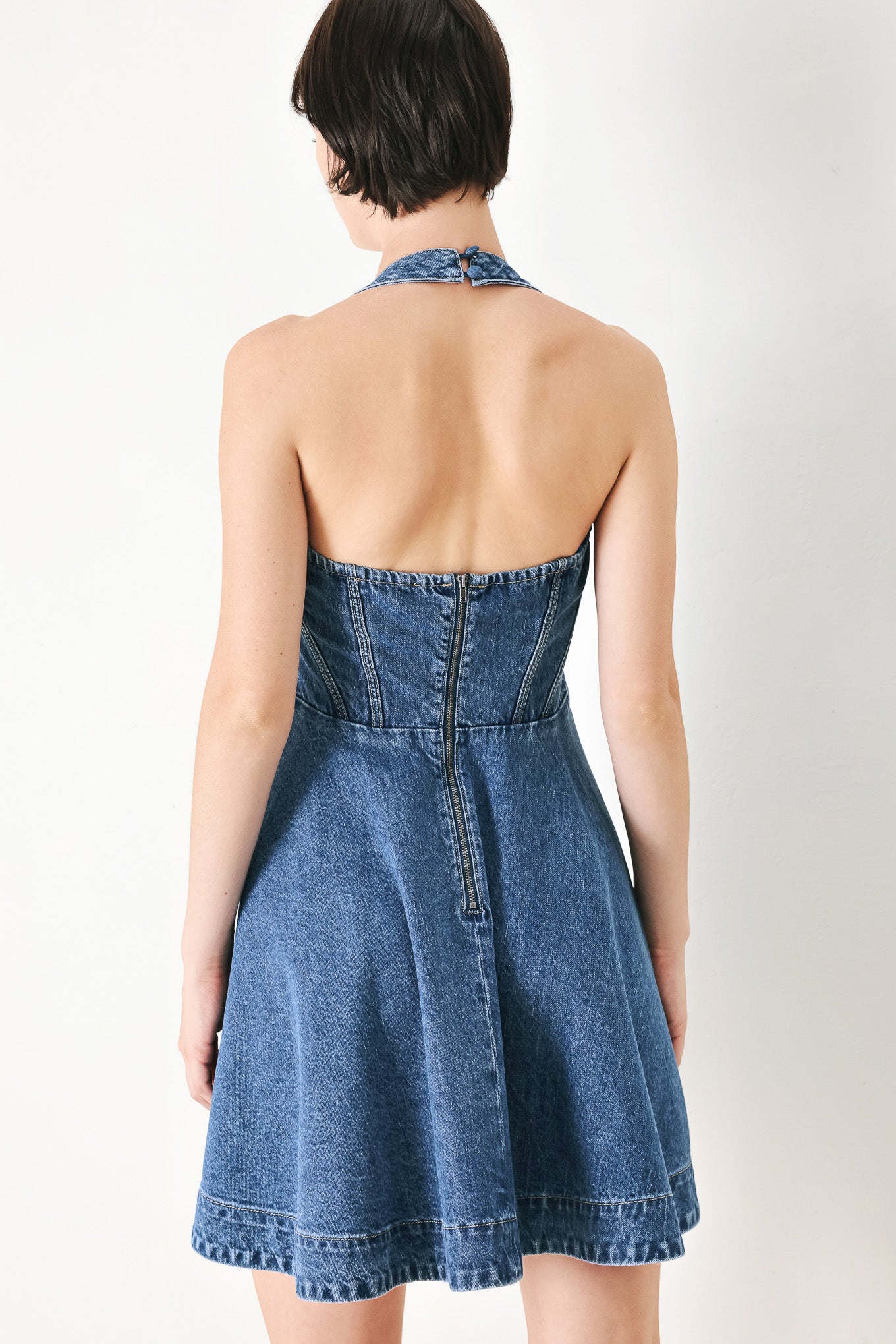 A washed denim mini dress featuring halter neckline with back button closure, full circle skirt bare back with zipper closure