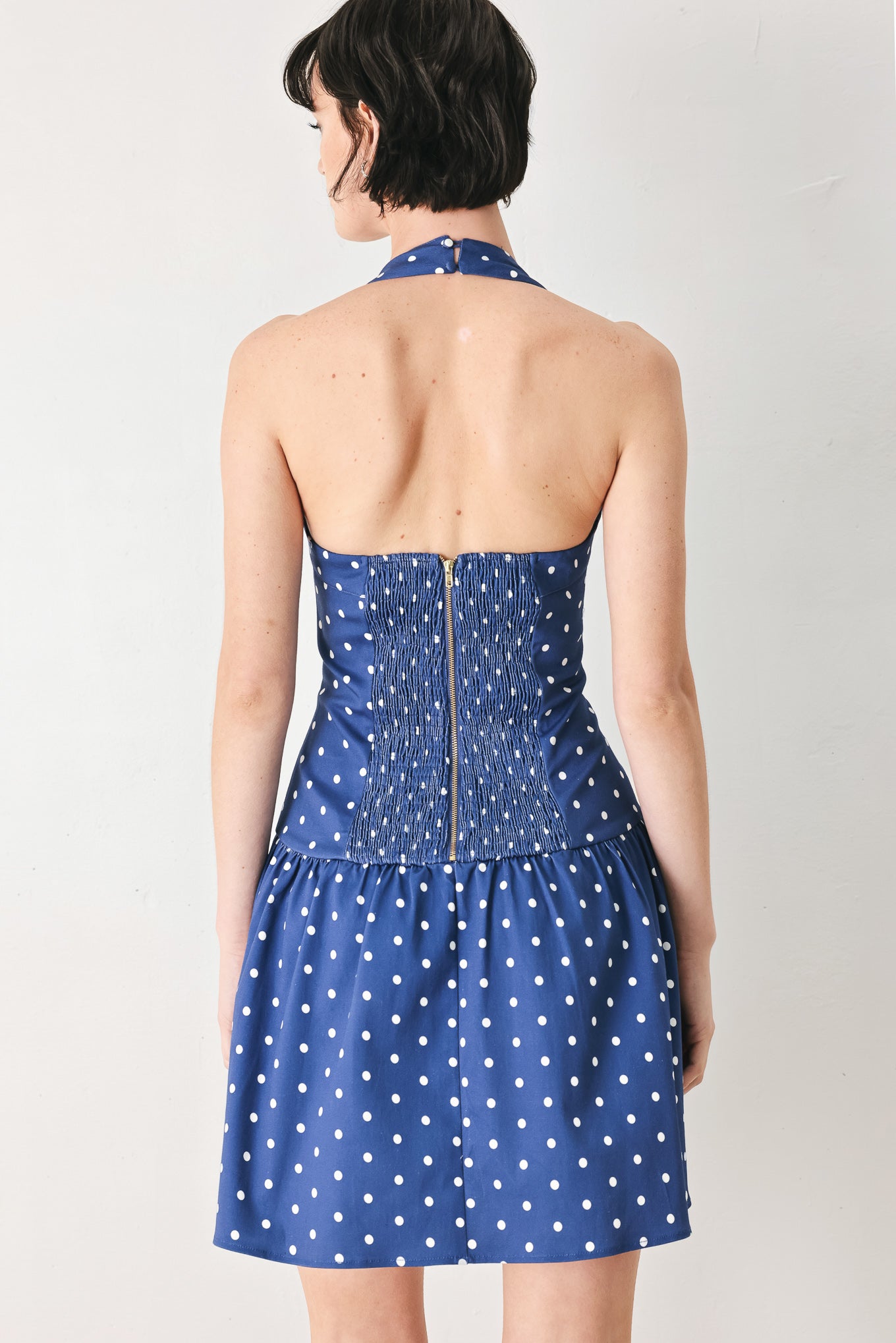 Woman wearing a blue polka dot mini dress featuring straight neckline with halter strap, drop waist and bare back with smocking zipper closure | FREEMARKET