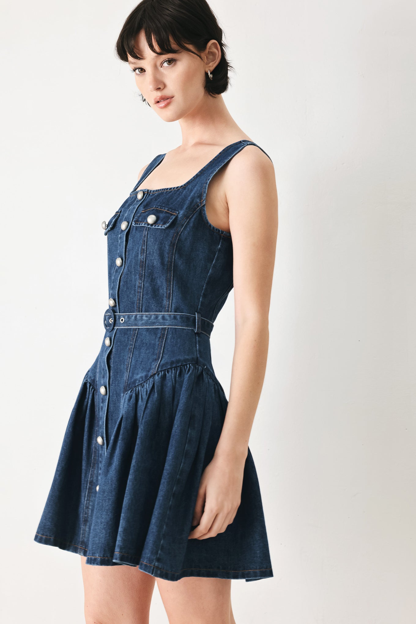 Woman wearing a blue denim sleeveless mini dress featuring square neckline, front button closure, self belt and side shirred full skirt | FREEMARKET