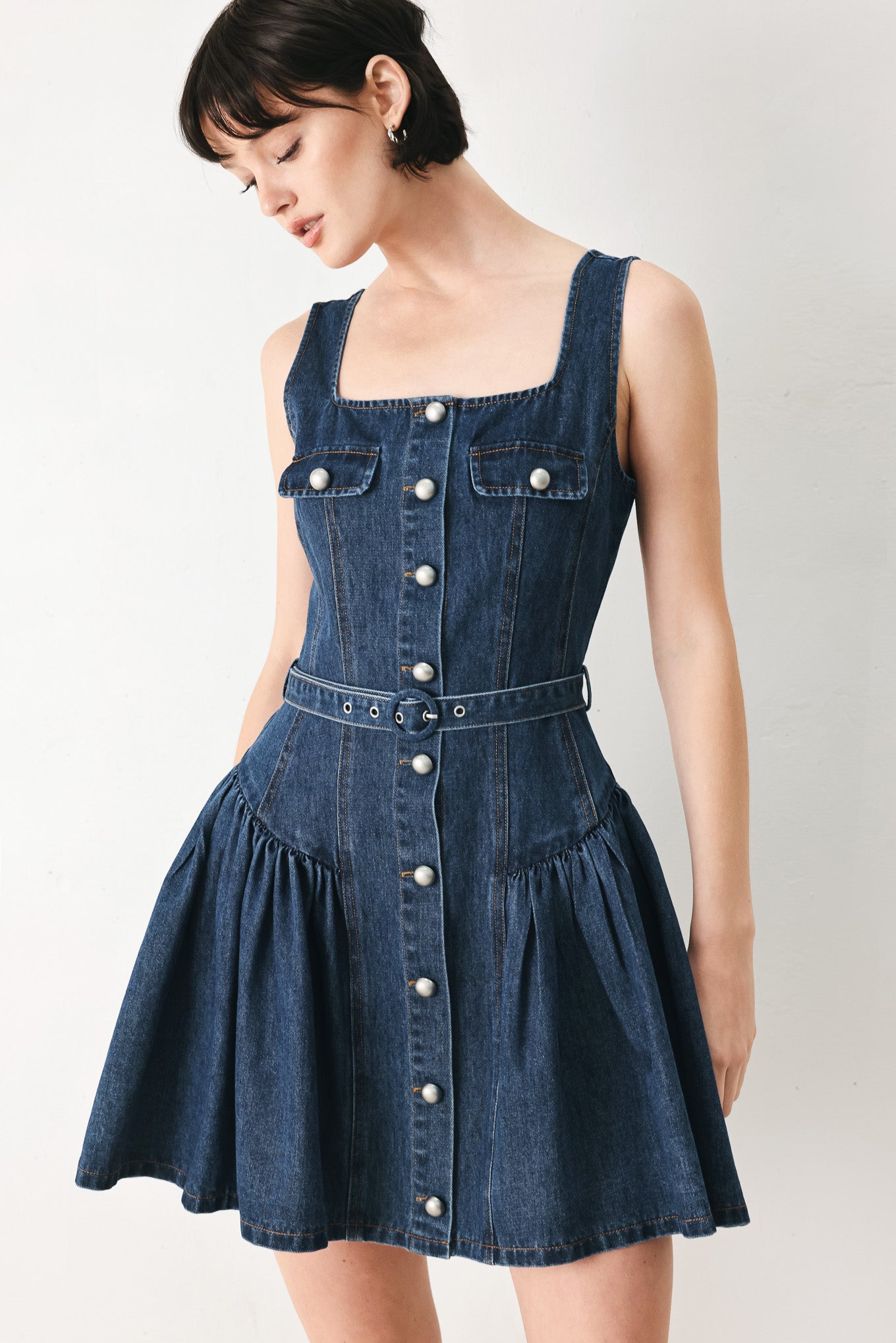 Woman wearing a blue denim sleeveless mini dress featuring square neckline, front button closure, self belt and side shirred full skirt | FREEMARKET