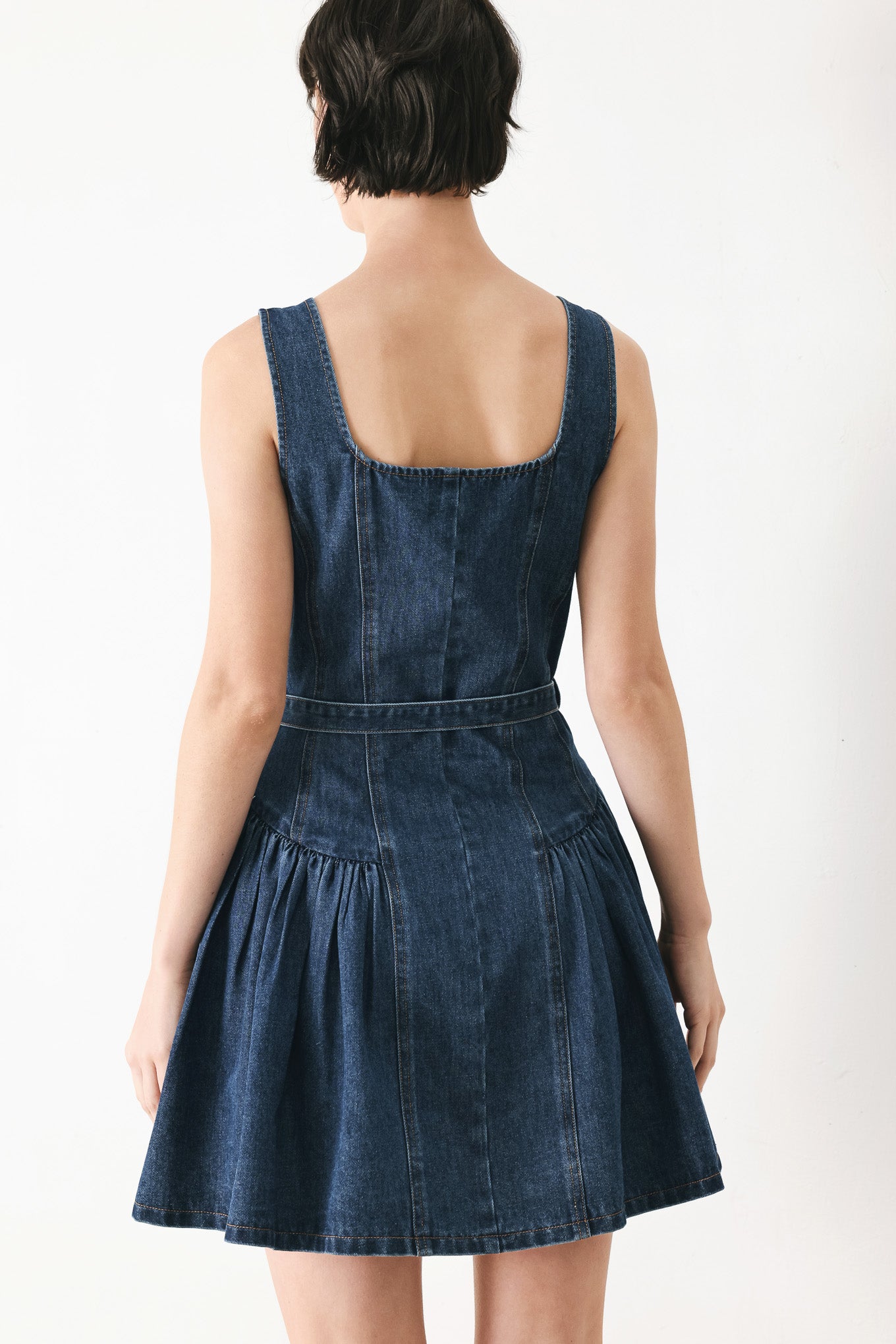 Woman wearing a blue denim sleeveless mini dress featuring square neckline, front button closure, self belt and side shirred full skirt | FREEMARKET
