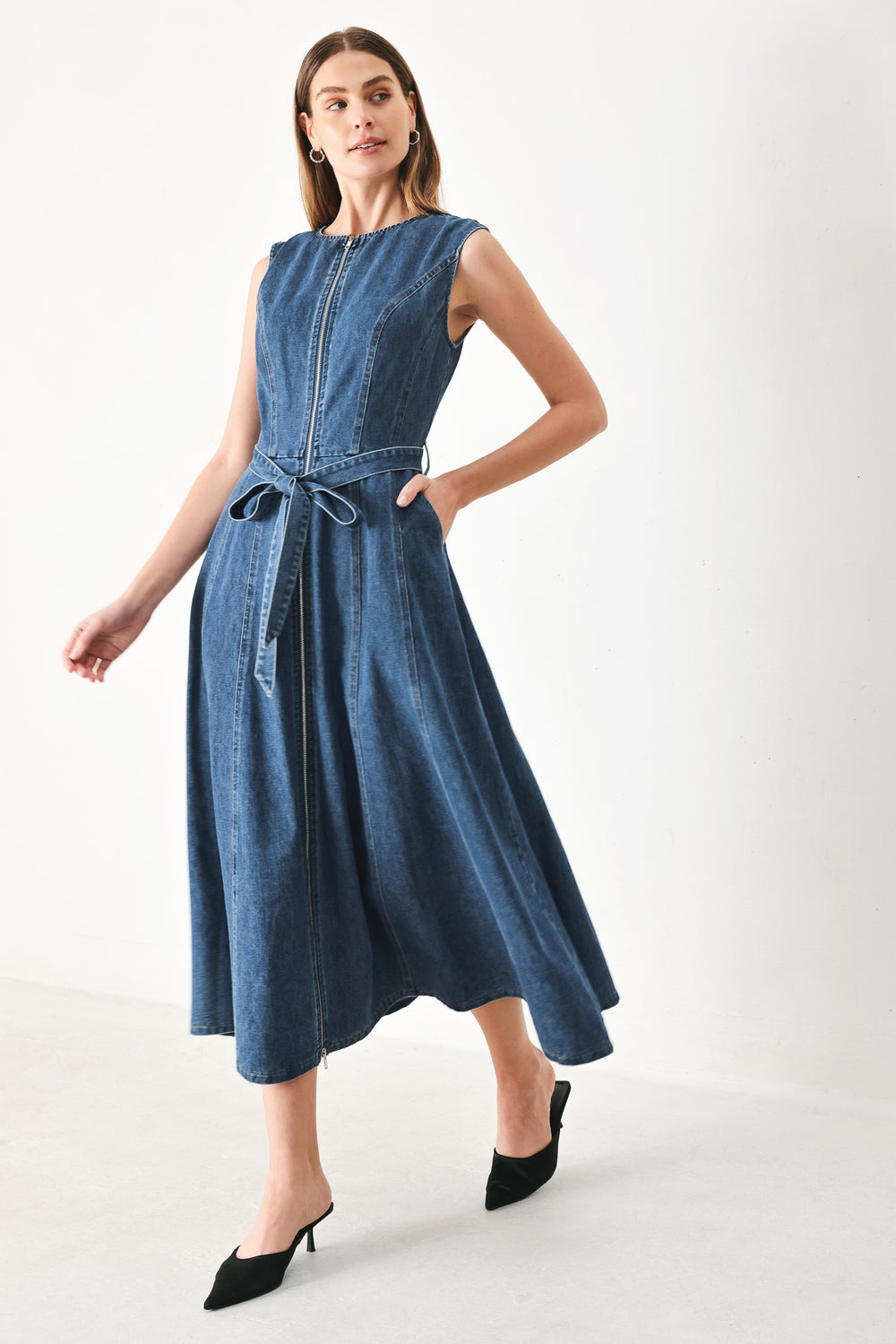 Woman wearing a blue denim midi dress featuring round neckline, sleeveless, self sash tie, circle skirt, side pockets and front zipper closure | FREEMARKET | Flying Tomato