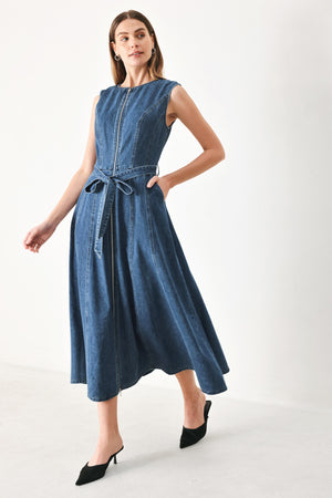 Woman wearing a blue denim midi dress featuring round neckline, sleeveless, self sash tie, circle skirt, side pockets and front zipper closure | FREEMARKET | Flying Tomato