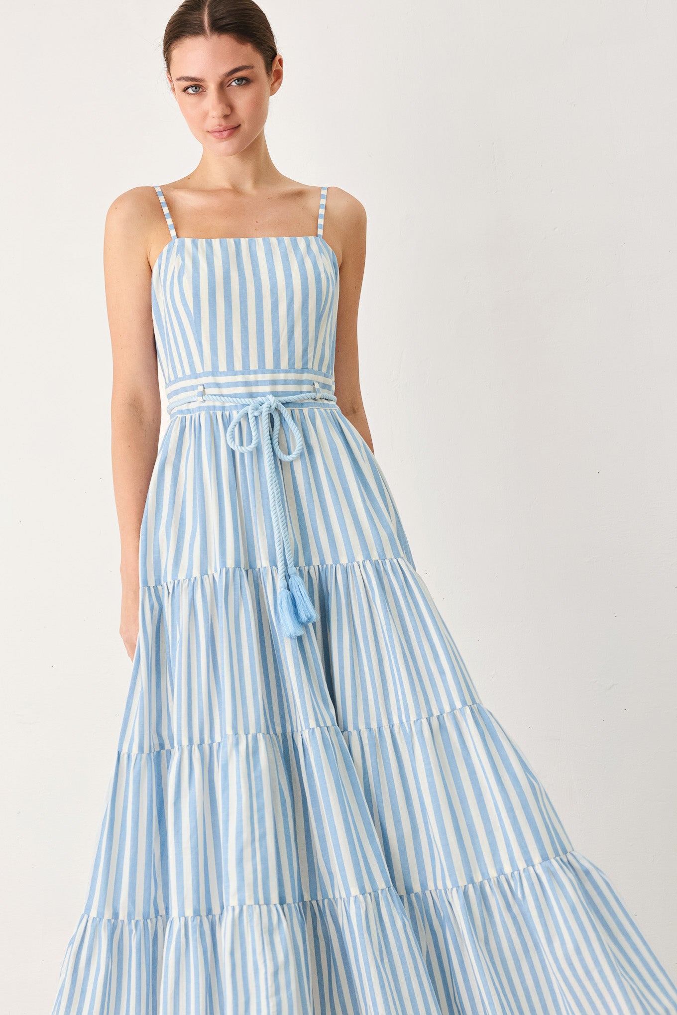 A woman wearing a light blue and white vertically striped maxi dress with thin shoulder straps, a matching tie belt at the waist, and a tiered skirt, photographed against a plain light background.
