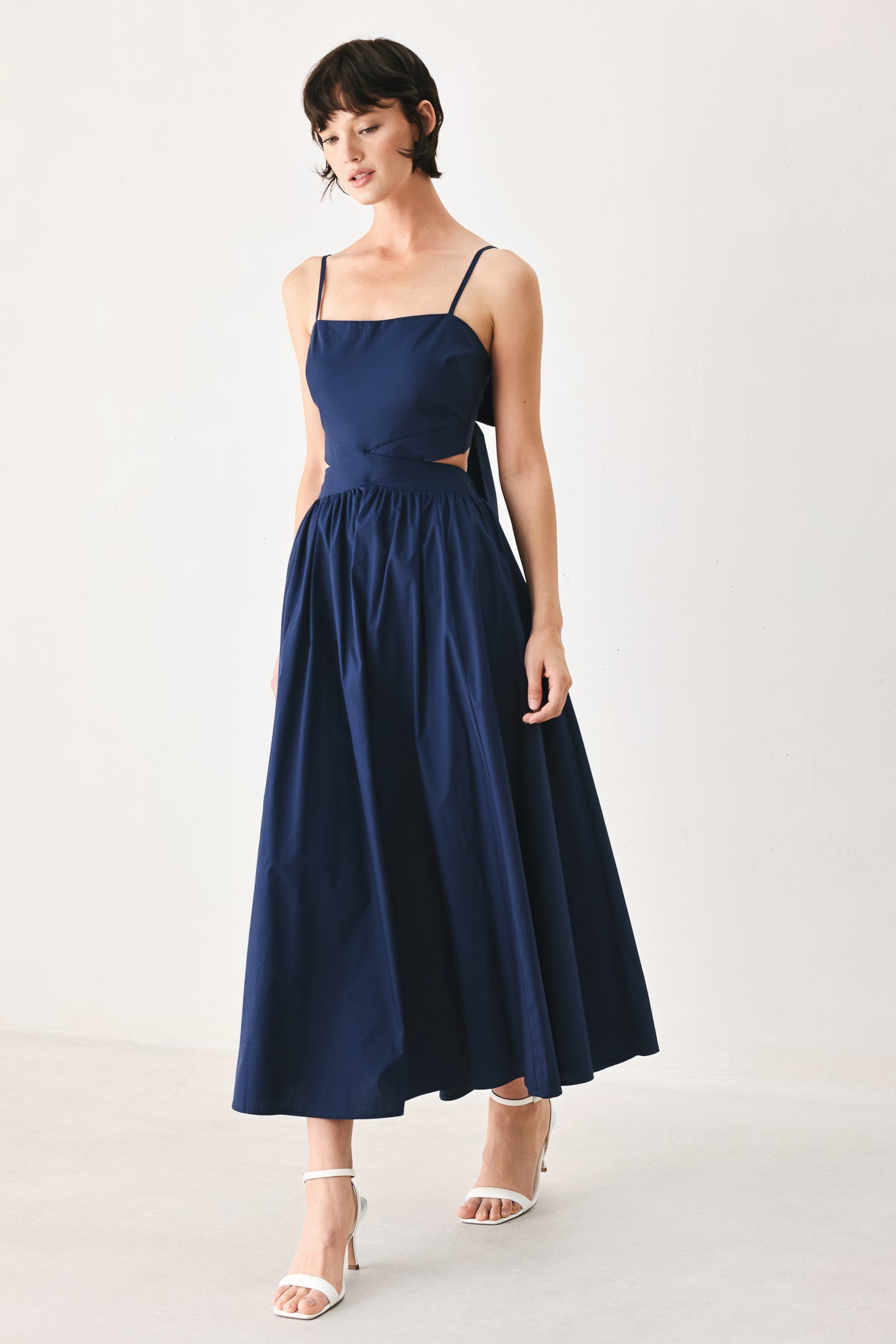 Woman wearing a navy midi dress featuring straight neckline, straps, waist cutout, full skirt, and tie detail | FREEMARKET
