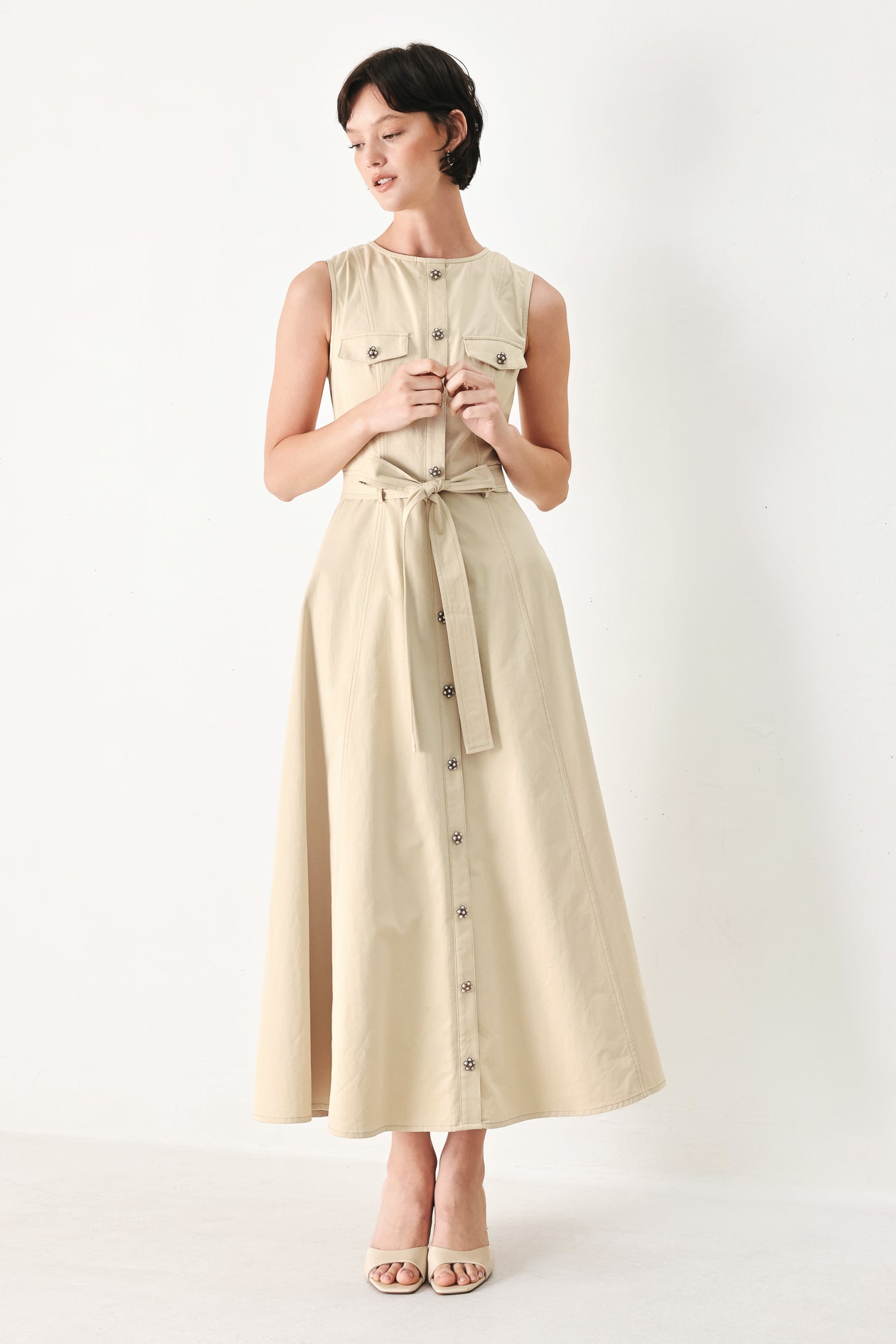 Woman wearing a beige khaki sleeveless midi dress with round neckline, front button detail, self sash tie, circle skirt and back zipper closure | FREEMARKET