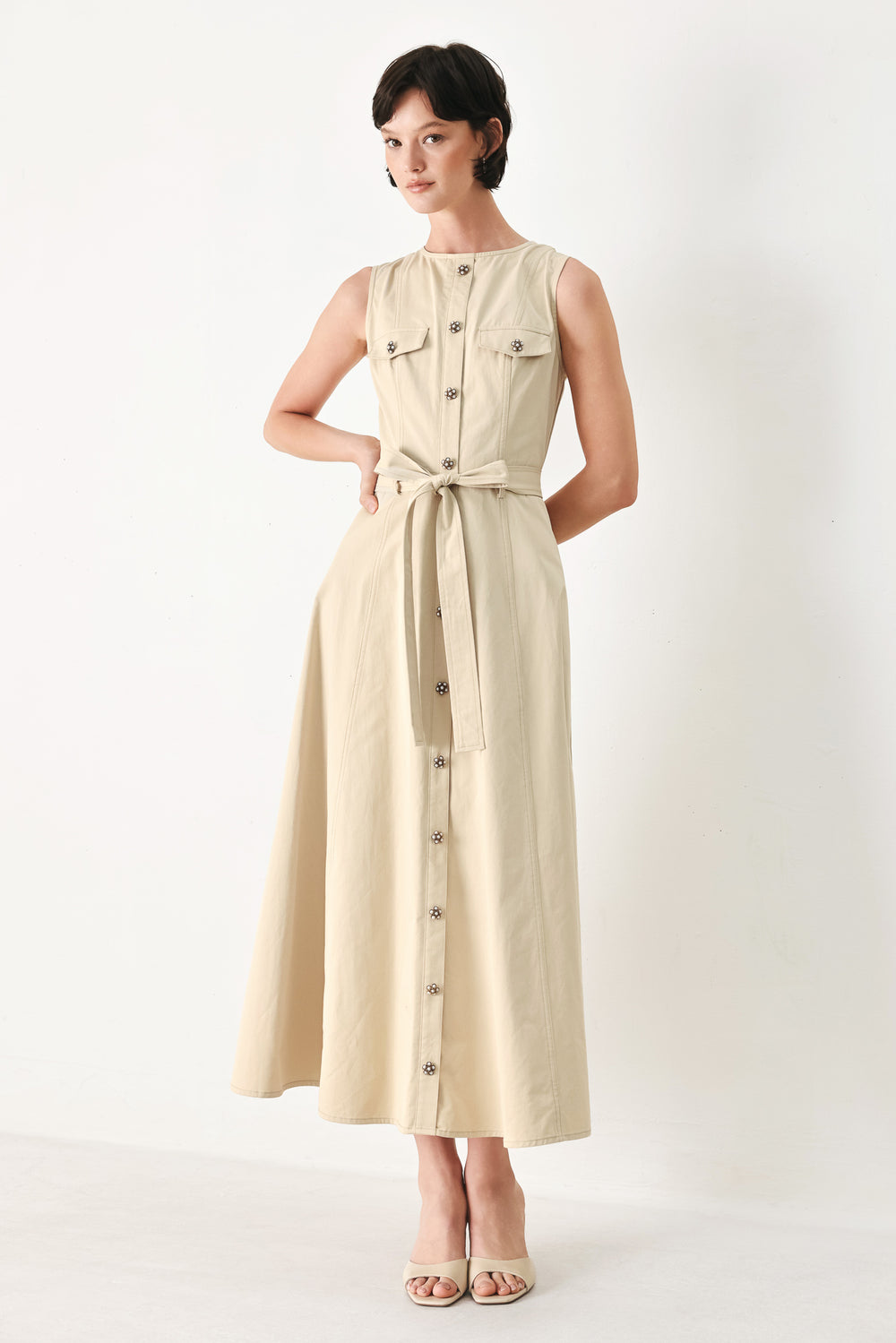 Woman wearing a beige khaki sleeveless midi dress with round neckline, front button detail, self sash tie, circle skirt and back zipper closure | FREEMARKET