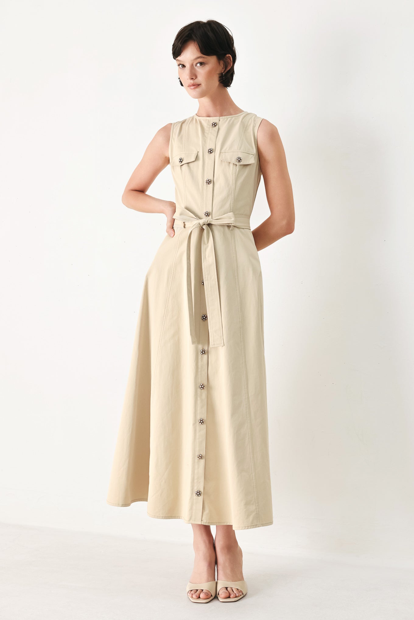 Woman wearing a beige khaki sleeveless midi dress with round neckline, front button detail, self sash tie, circle skirt and back zipper closure | FREEMARKET