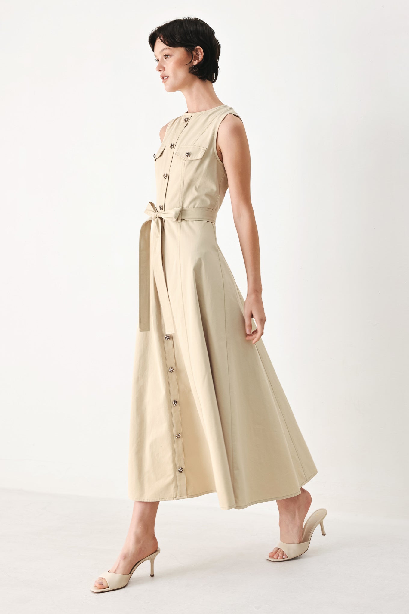 Woman wearing a beige khaki sleeveless midi dress with round neckline, front button detail, self sash tie, circle skirt and back zipper closure | FREEMARKET