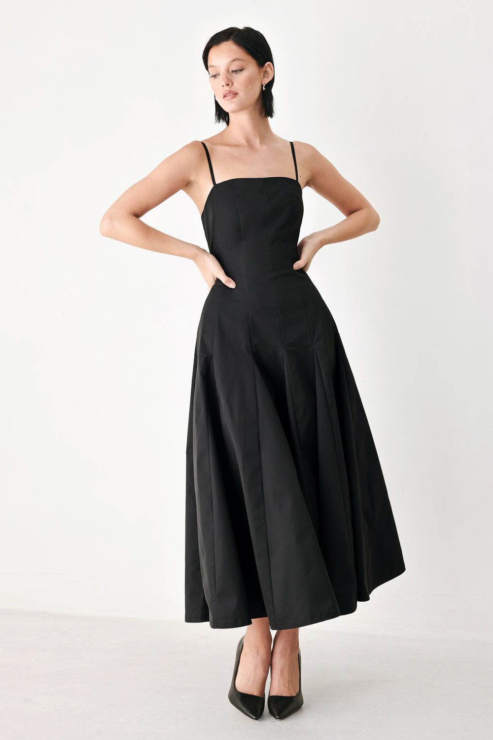 A woman modeling a black midi dress with thin spaghetti straps, a structured square neckline, and a flared skirt | FREEMARKET