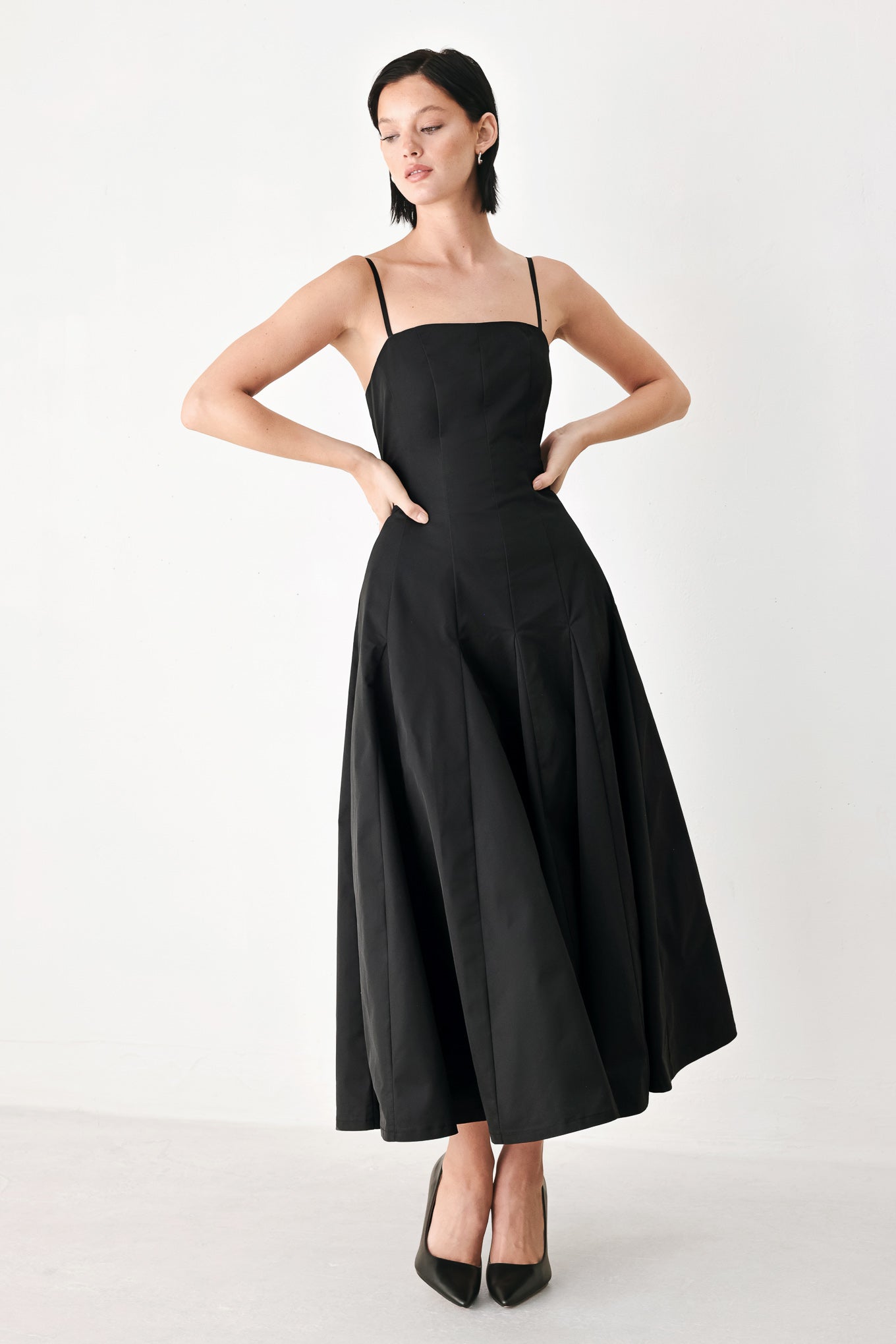 A woman modeling a black midi dress with thin spaghetti straps, a structured square neckline, and a flared skirt | FREEMARKET
