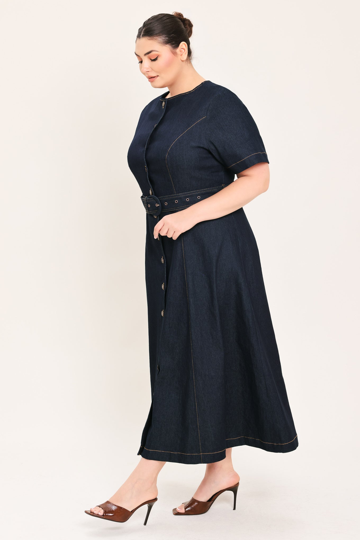A woman modeling a dark indigo denim midi dress with short sleeves, a button-down front, and a matching belt with a metal buckle | Plus Curve Flying Tomato