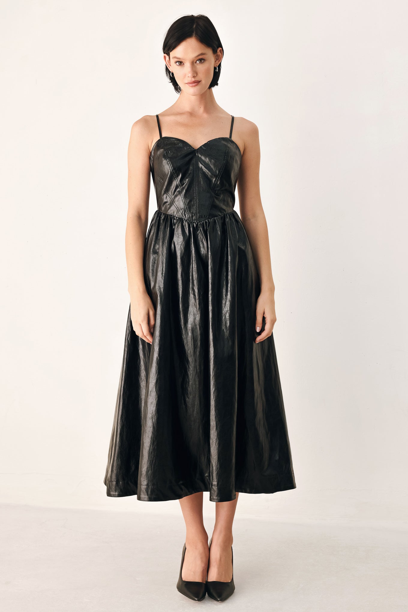 A woman modeling a black faux leather midi dress with a shiny, textured finish, featuring thin spaghetti straps, a sweetheart neckline, a gathered corset-style bodice, and a flared skirt | FREEMARKET