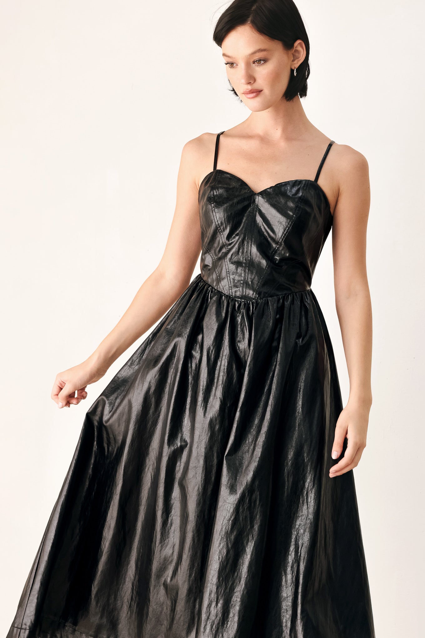 A woman modeling a black faux leather midi dress with a shiny, textured finish, featuring thin spaghetti straps, a sweetheart neckline, a gathered corset-style bodice, and a flared skirt | FREEMARKET