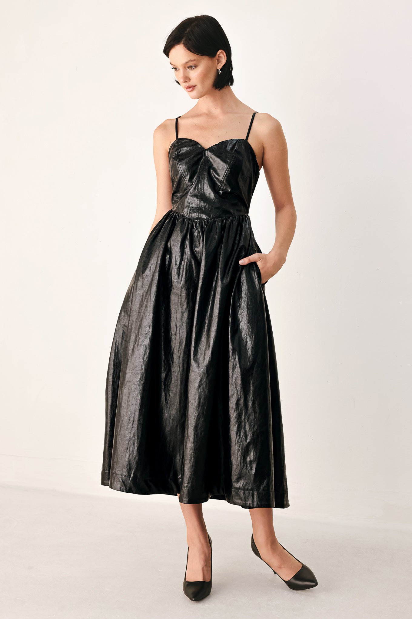 A woman modeling a black faux leather midi dress with a shiny, textured finish, featuring thin spaghetti straps, a sweetheart neckline, a gathered corset-style bodice, and a flared skirt | FREEMARKET