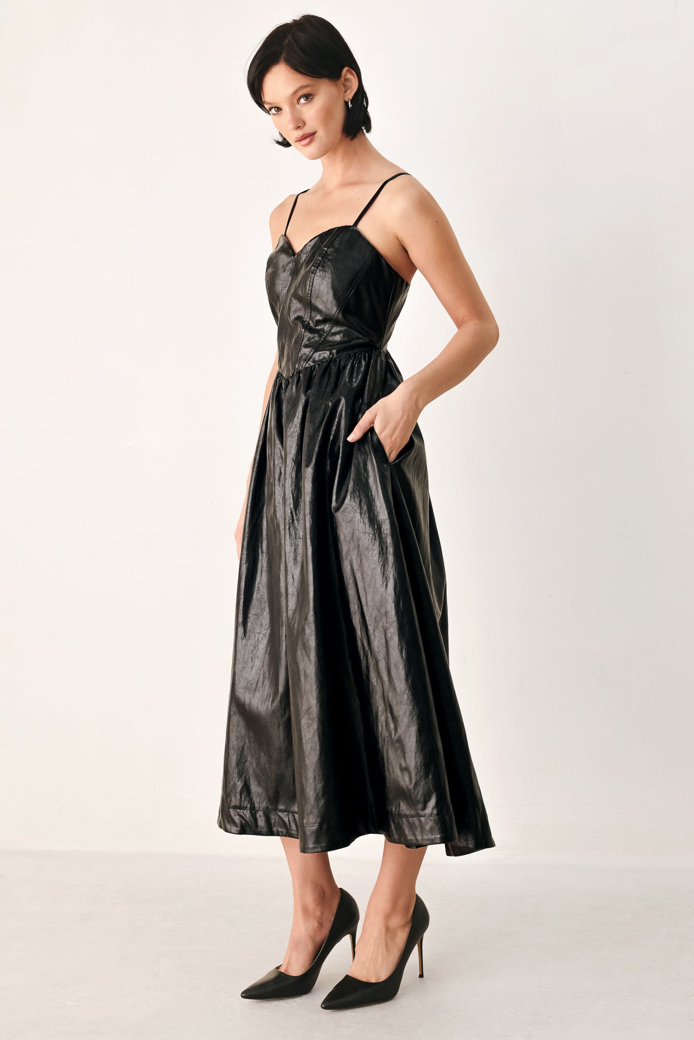 A woman modeling a black faux leather midi dress with a shiny, textured finish, featuring thin spaghetti straps, a sweetheart neckline, a gathered corset-style bodice, and a flared skirt | FREEMARKET