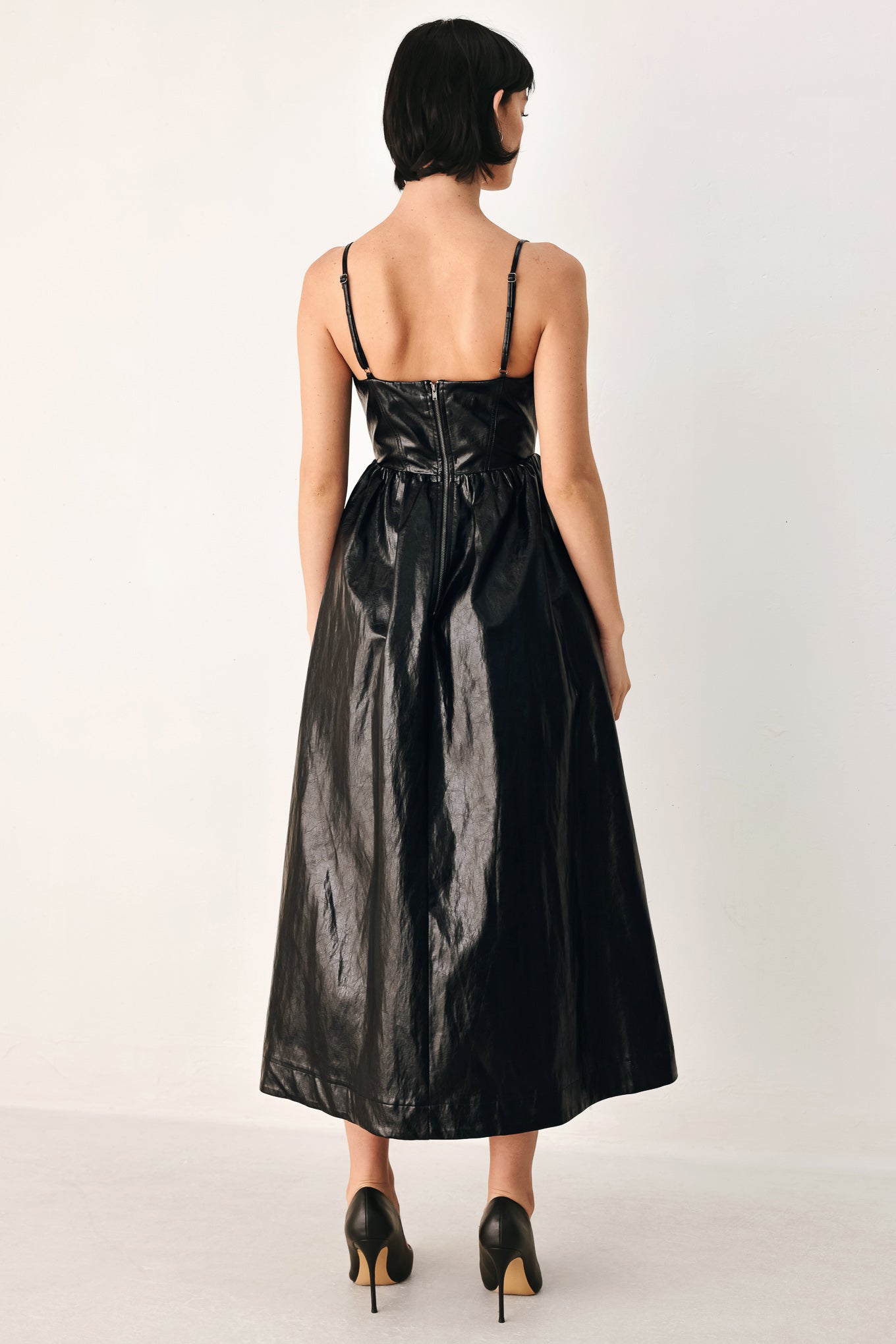 A woman modeling a black faux leather midi dress with a shiny, textured finish, featuring thin spaghetti straps, a sweetheart neckline, a gathered corset-style bodice, and a flared skirt | FREEMARKET