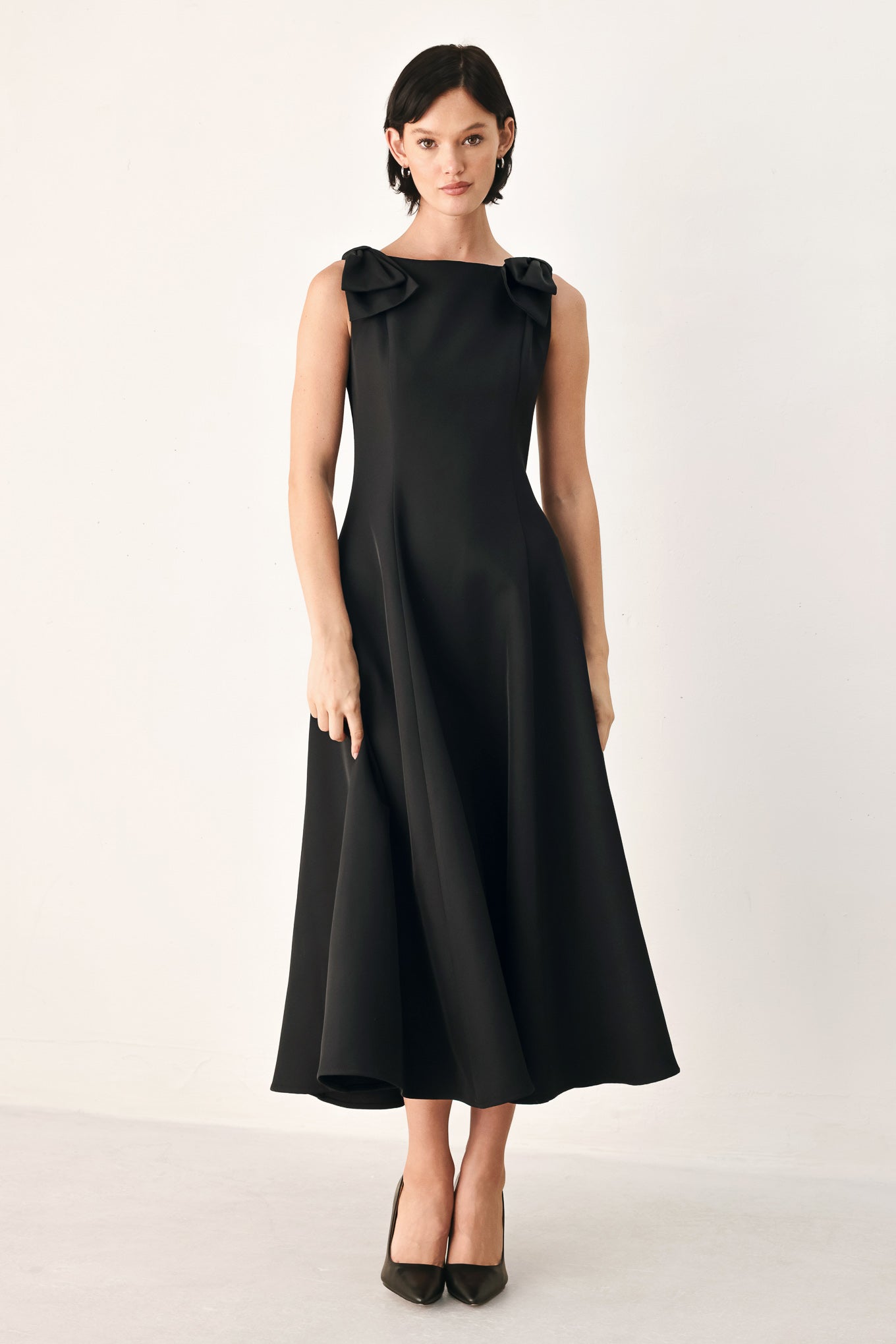 A woman modeling a sleeveless black midi dress with a boat neckline and large bow details on the shoulders | FREEMARKET