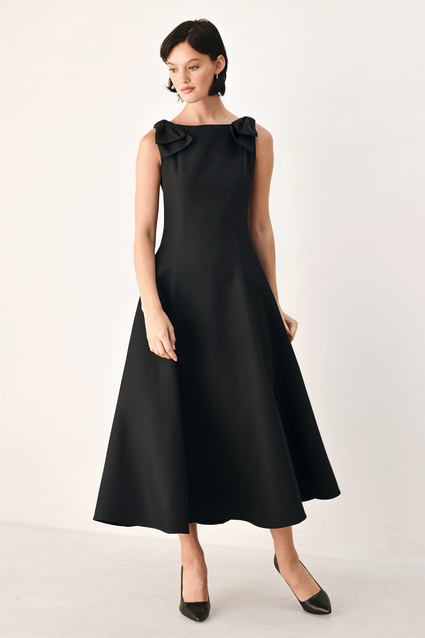 A woman modeling a sleeveless black midi dress with a boat neckline and large bow details on the shoulders | FREEMARKET