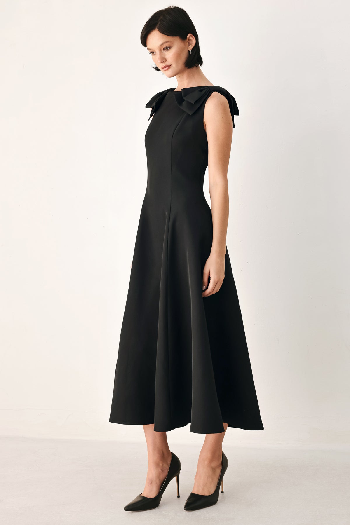 A woman modeling a sleeveless black midi dress with a boat neckline and large bow details on the shoulders | FREEMARKET