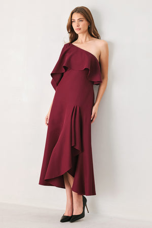 Woman wearing a burgundy midi dress with with an asymmetrical neckline with a large ruffle and a ruffle along the leg slit | FREEMARKET