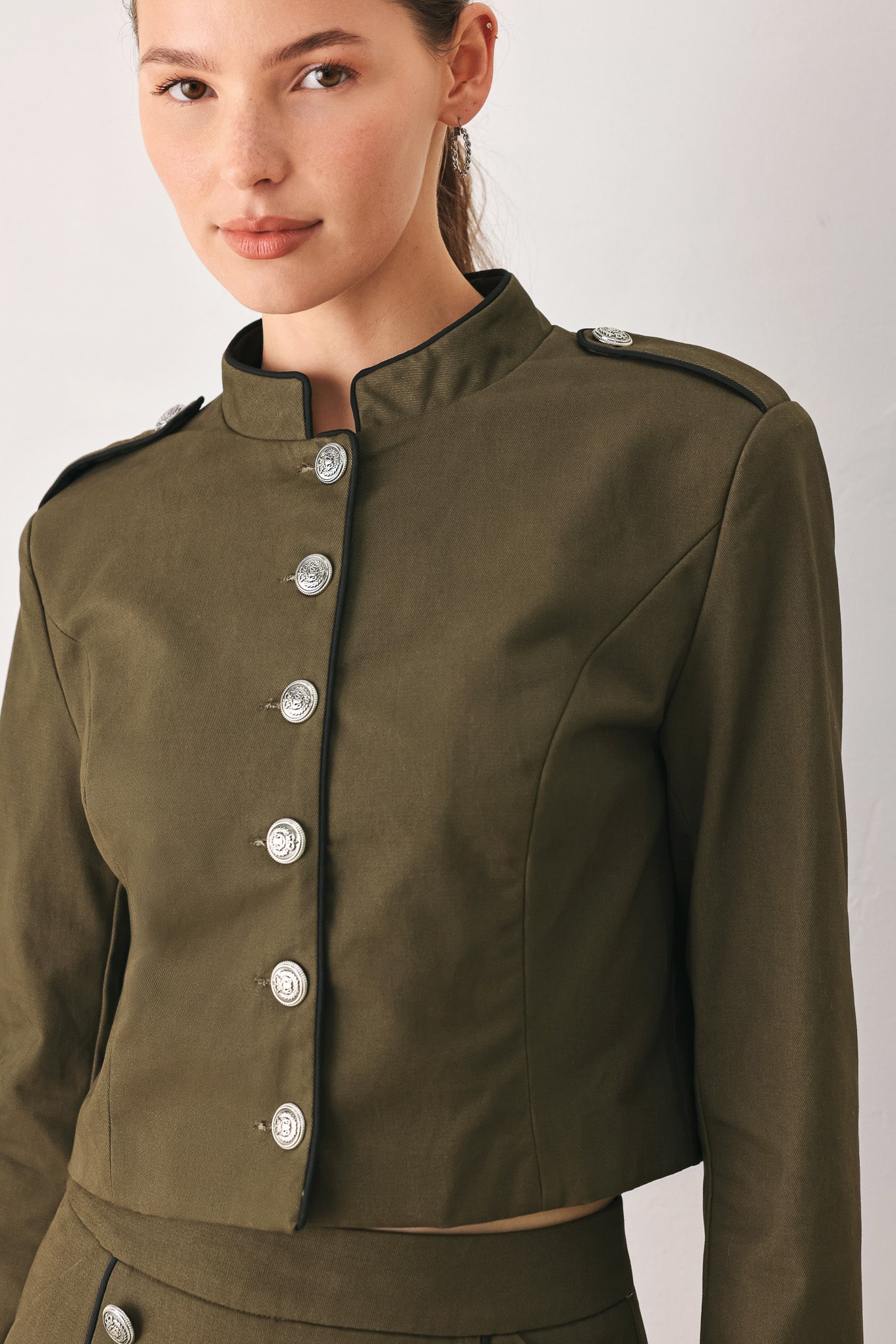 A close-up of a model wearing an olive green cropped jacket with a high stand collar, silver buttons down the front, and shoulder epaulets | FREEMARKET