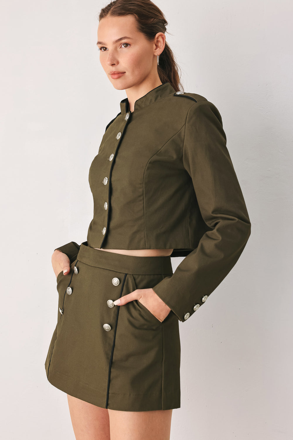 A close-up of a model wearing an olive green cropped jacket with a high stand collar, silver buttons down the front, and shoulder epaulets | FREEMARKET
