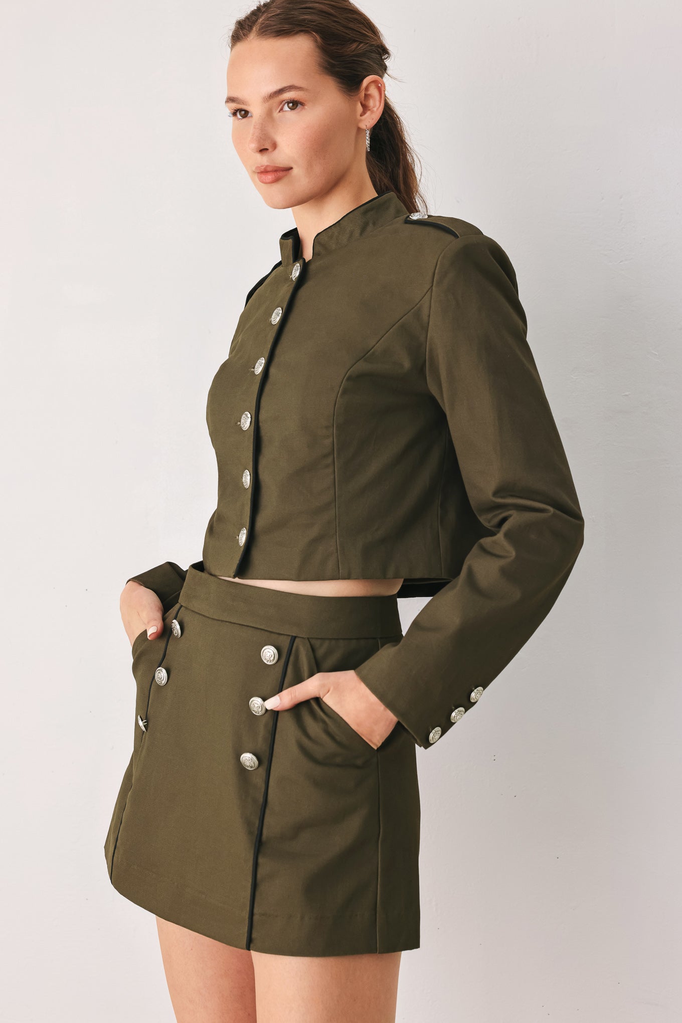 A close-up of a model wearing an olive green cropped jacket with a high stand collar, silver buttons down the front, and shoulder epaulets | FREEMARKET