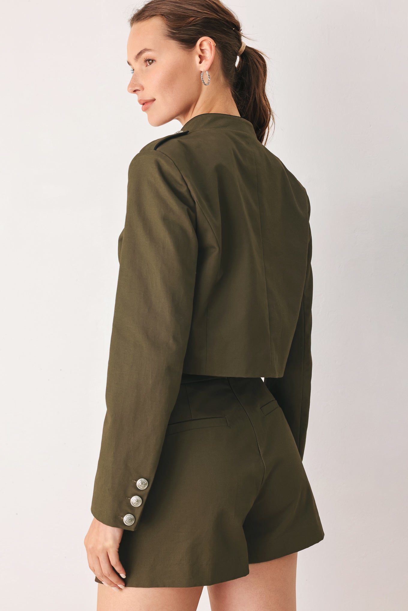 A close-up of a model wearing an olive green cropped jacket with a high stand collar, silver buttons down the front, and shoulder epaulets | FREEMARKET
