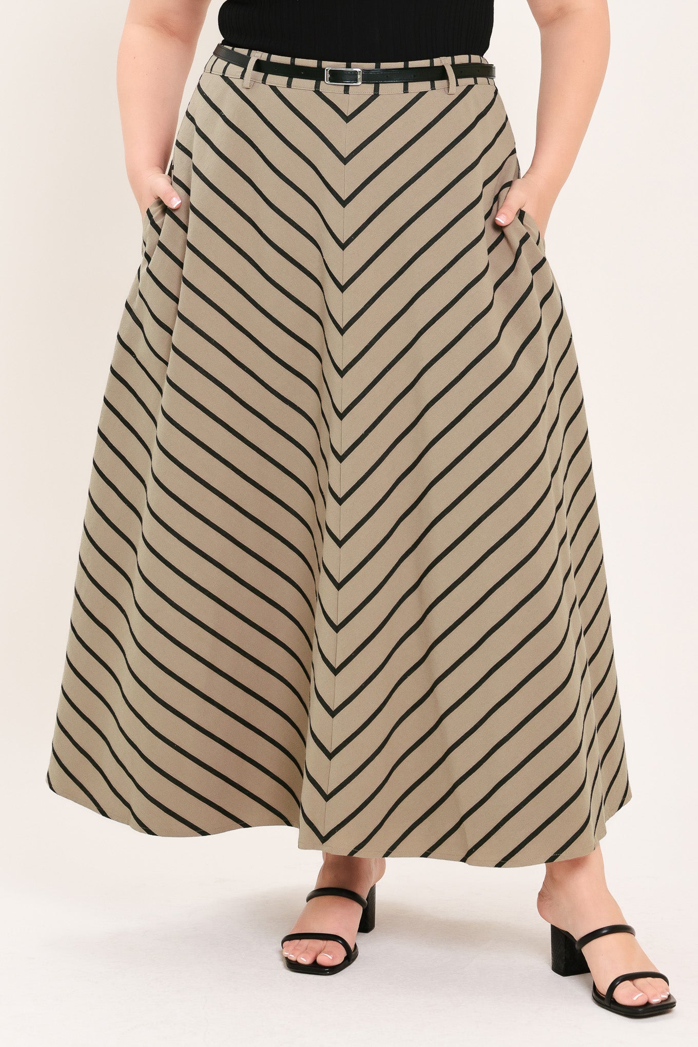 A woman wearing a beige A-line midi skirt with a black chevron stripe pattern, styled with a black belt, black top, and black heeled sandals | FREEMARKET Curve