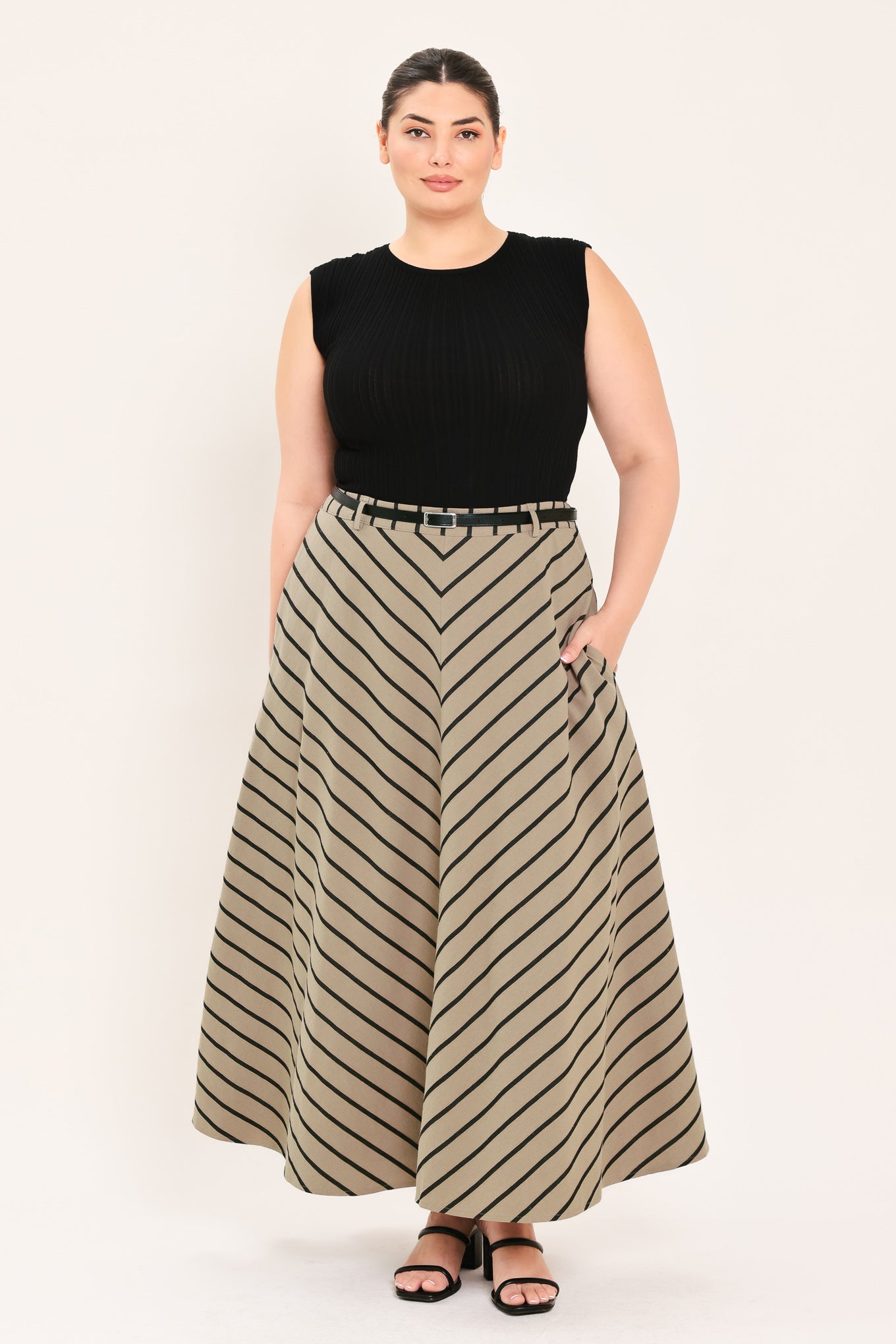 A woman wearing a beige A-line midi skirt with a black chevron stripe pattern, styled with a black belt, black top, and black heeled sandals | FREEMARKET Curve