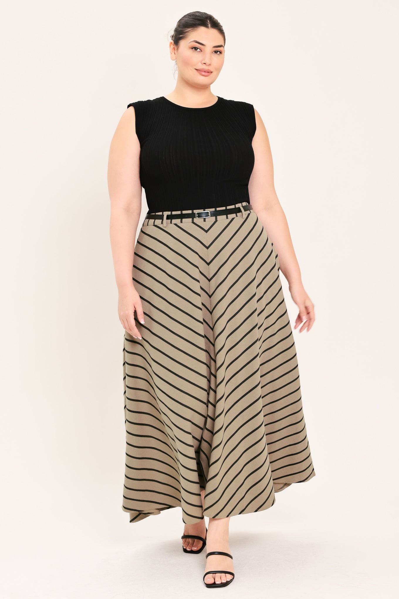 A woman wearing a beige A-line midi skirt with a black chevron stripe pattern, styled with a black belt, black top, and black heeled sandals | FREEMARKET Curve
