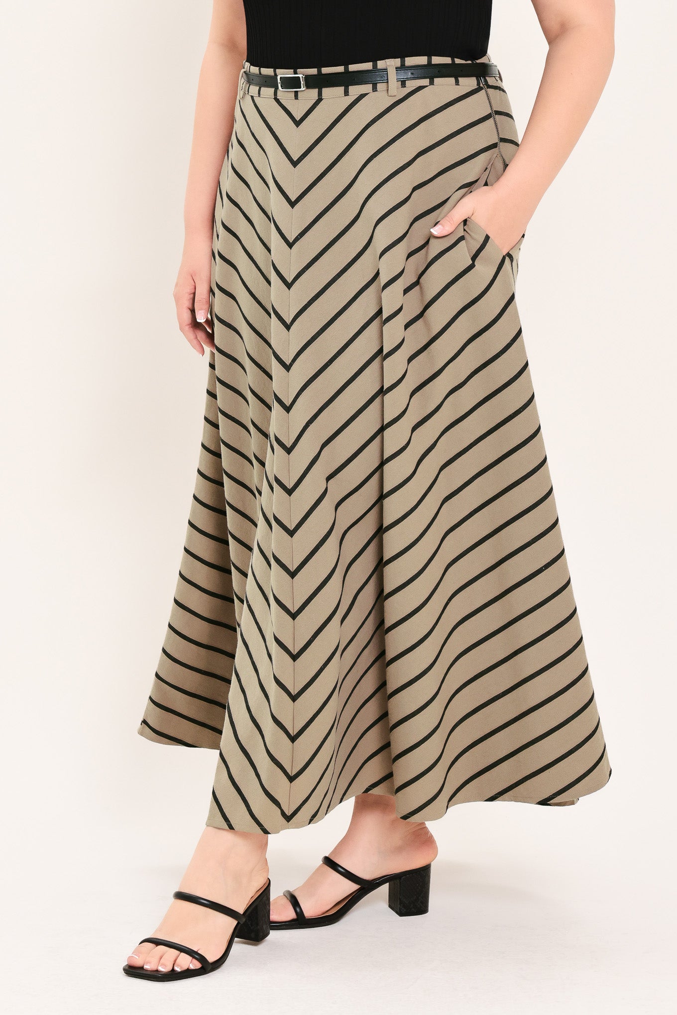 A woman wearing a beige A-line midi skirt with a black chevron stripe pattern, styled with a black belt, black top, and black heeled sandals | FREEMARKET Curve